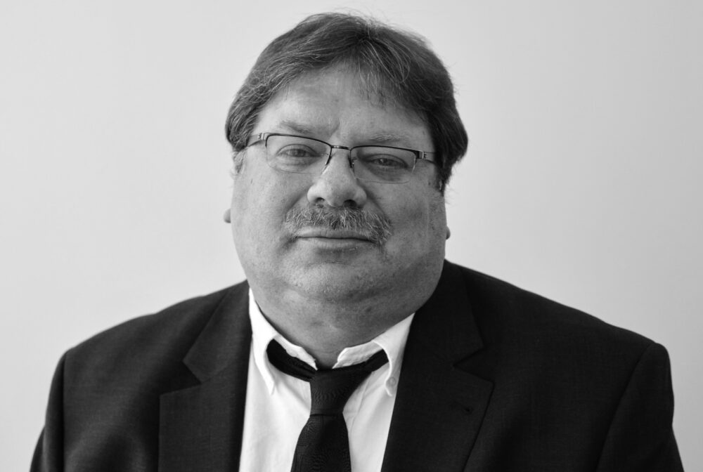 Darren wearing glasses and a suit, looking towards the camera in a neutral expression