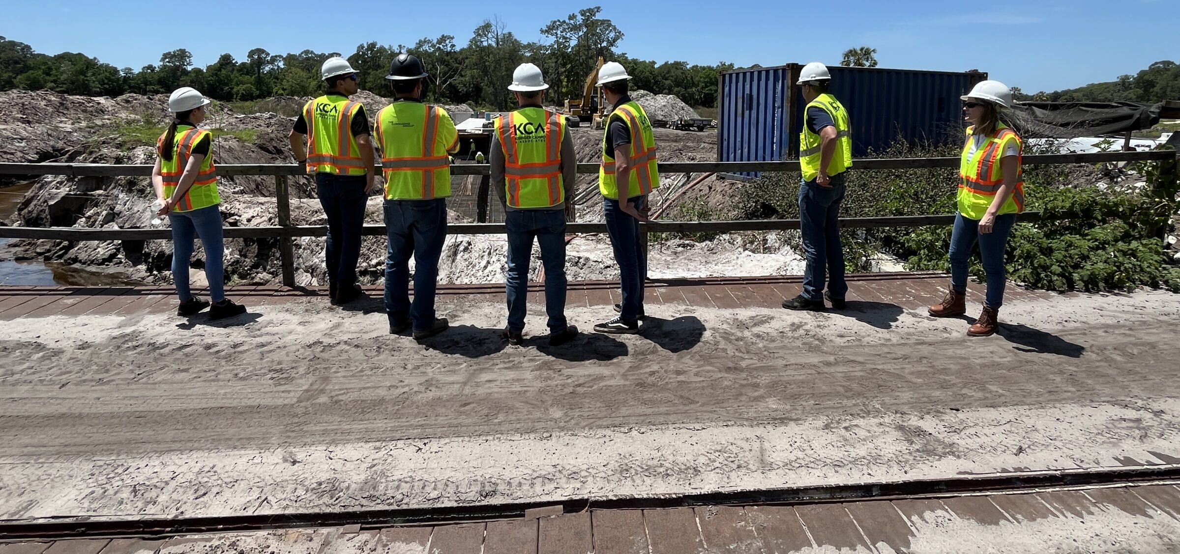 Seven construction workers in safety vests and hard hats discuss site plans