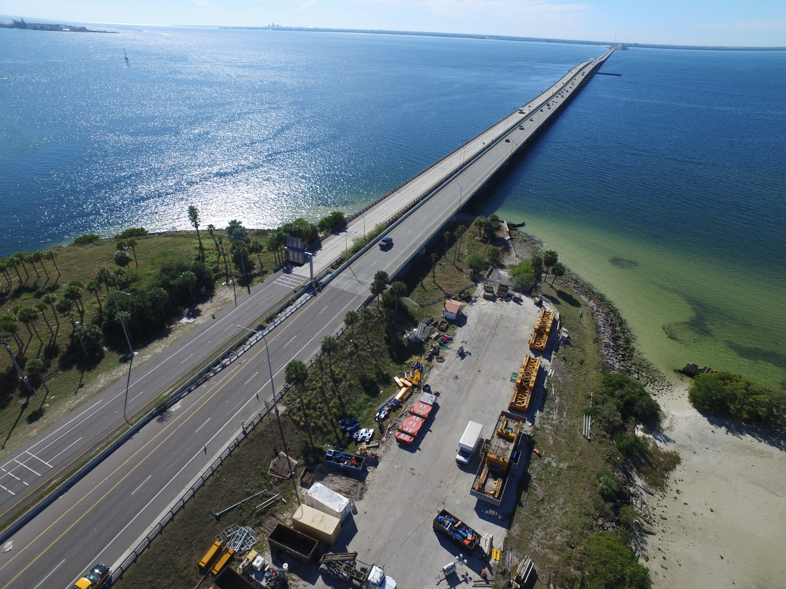 Aerial view of a long bridge over a large body of water, with lush greenery, parked construction vehicles, and equipment nearby
