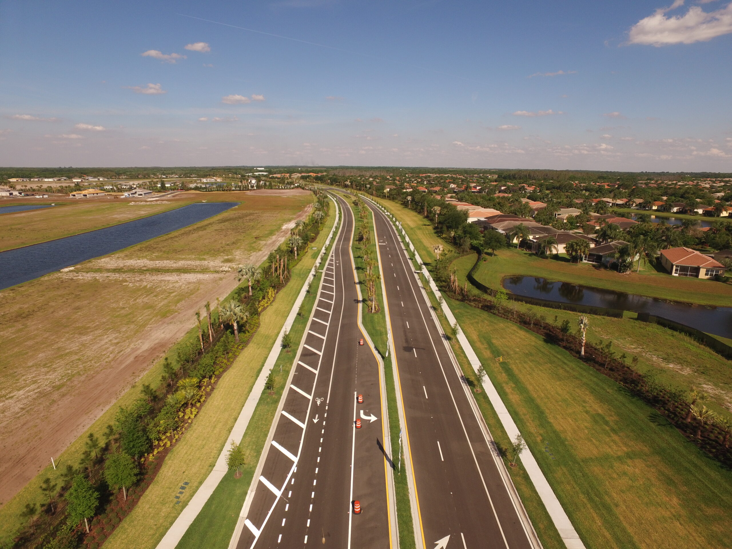 Aerial view of a wide road with surrounding greenery, houses, and water bodies under a clear blue sky