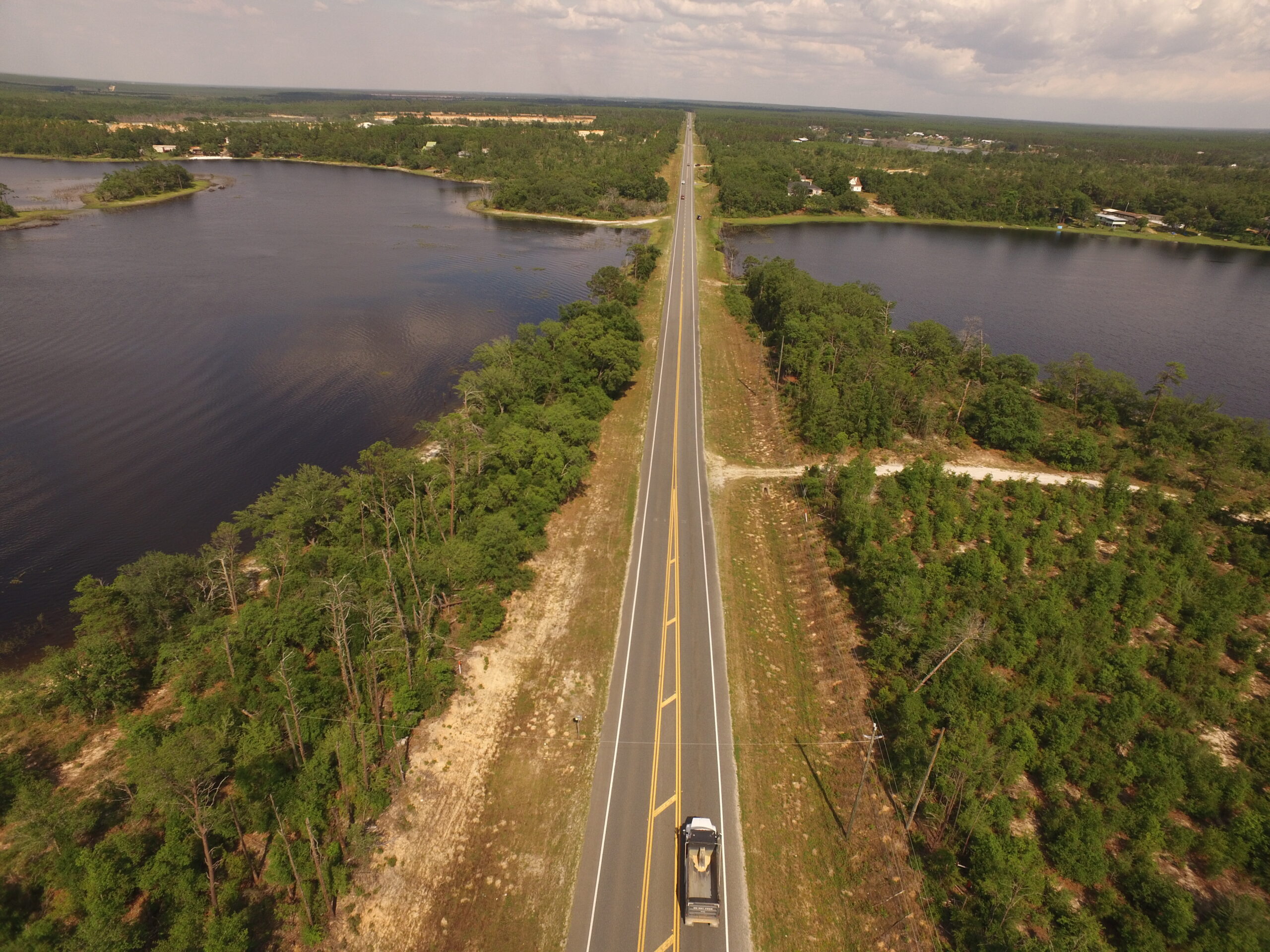 Aerial view of a long, straight road bordered by lakes and forests, with a truck driving on it