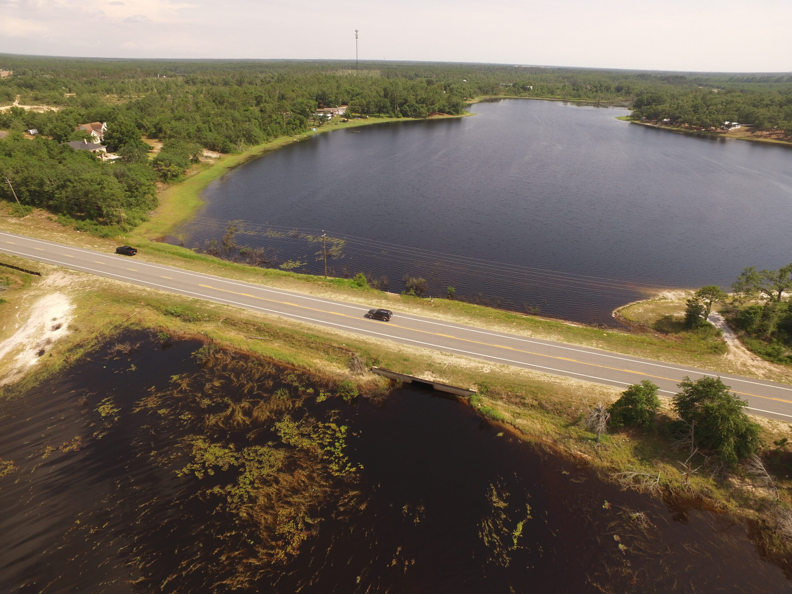 Aerial view of a tranquil lake bordered by lush green forest with a road and two black cars in the foreground