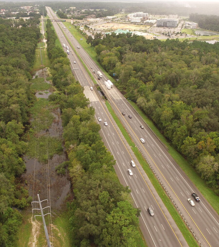 Aerial view of a highway surrounded by dense green forest and distant cityscape