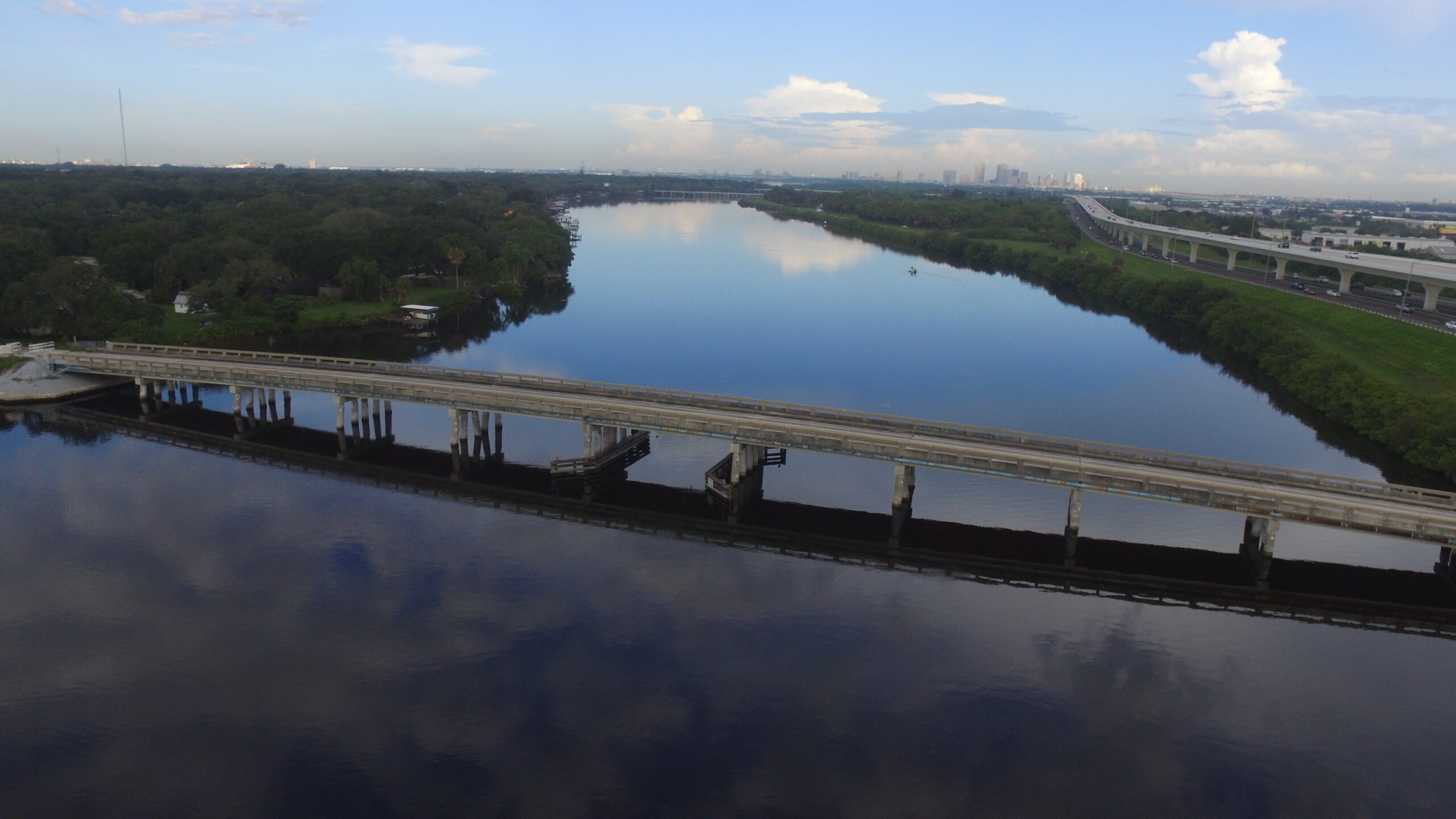 Aerial view of a wide river reflecting the sky, with a bridge crossing it. Green trees line the riverbanks, and a distant city skyline is visible against a partly cloudy sky