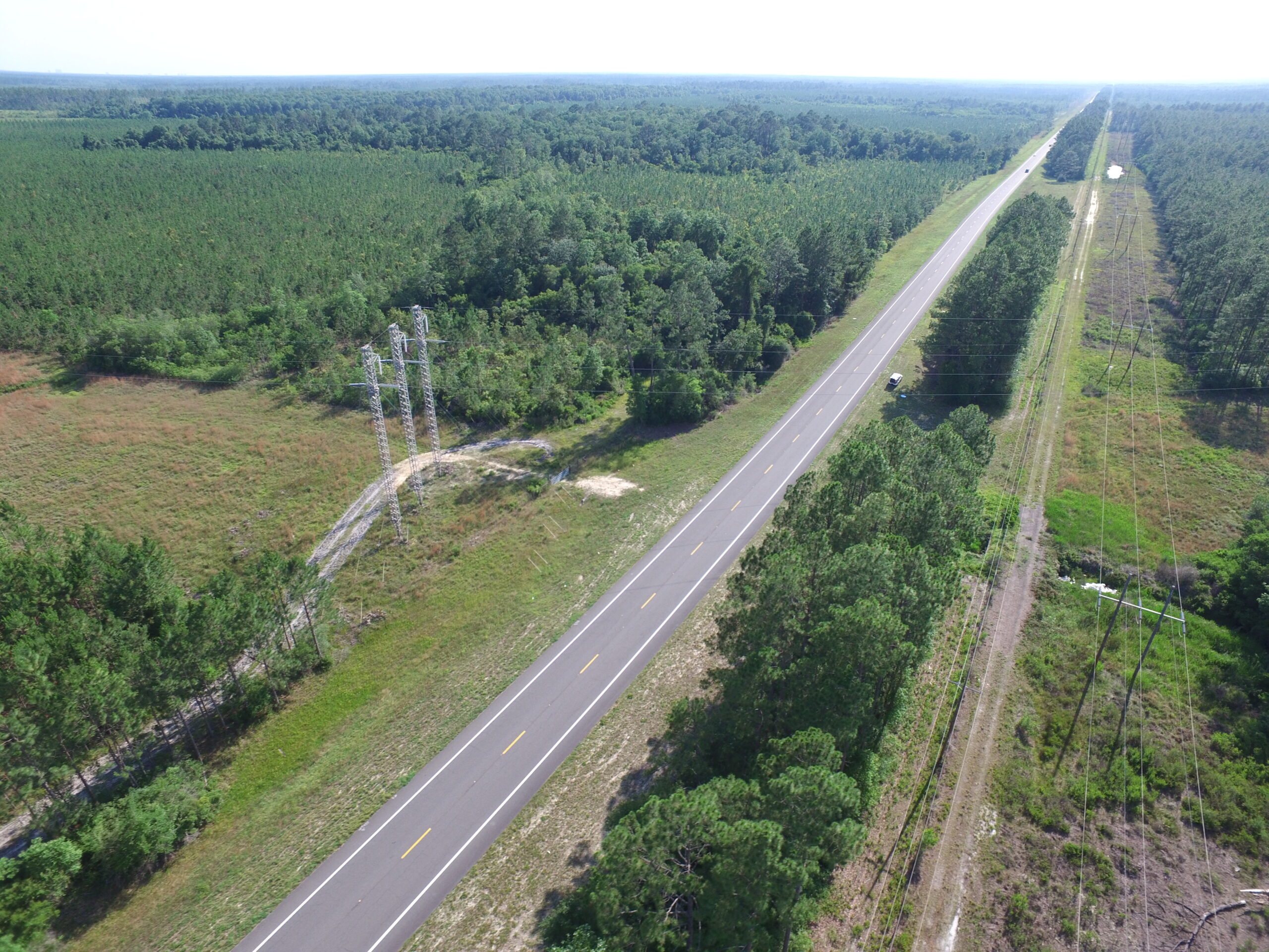 Aerial view of a straight road bordered by dense green forest and power lines