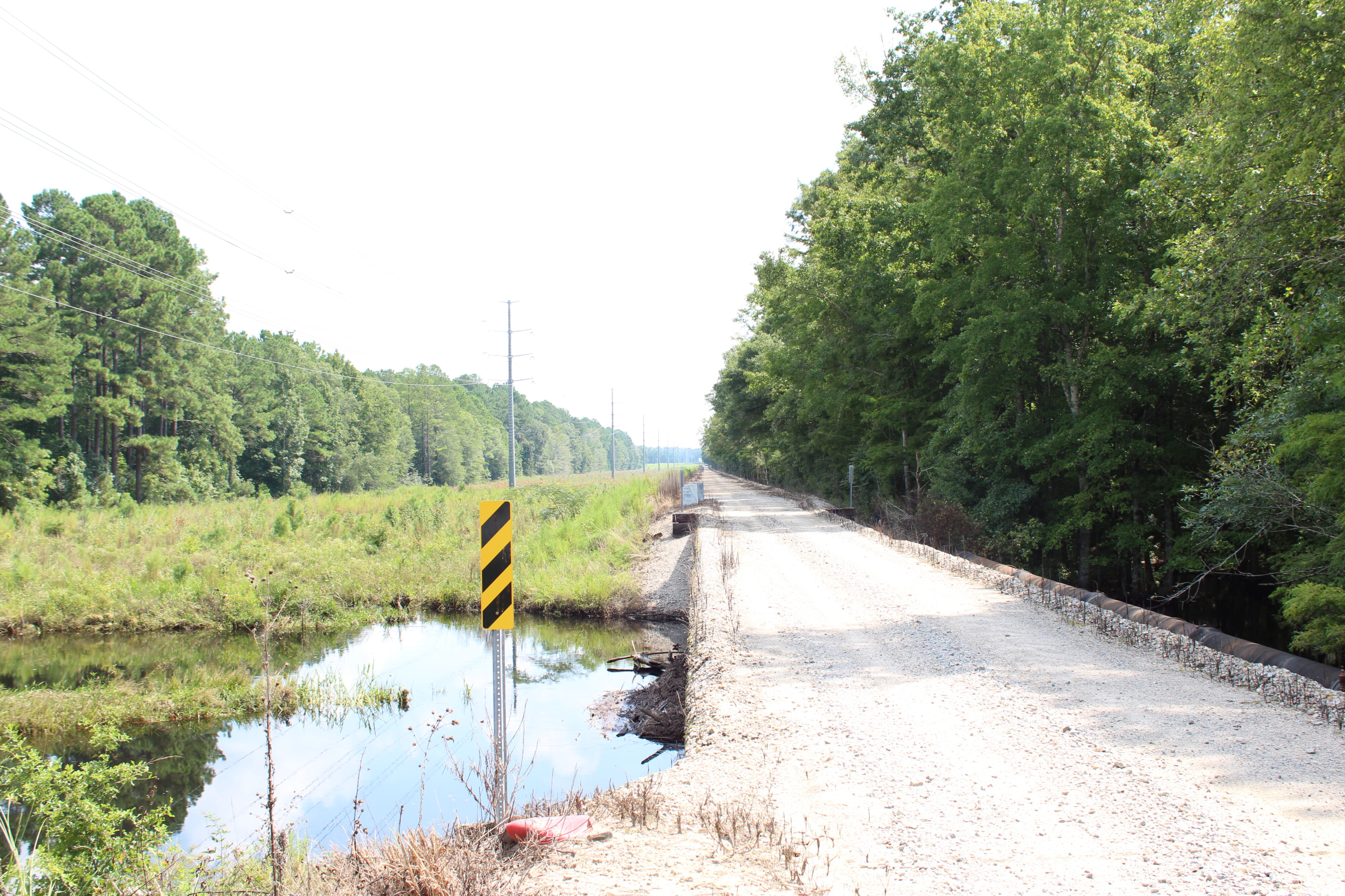 Gravel road beside forest and water, marked by striped caution sign