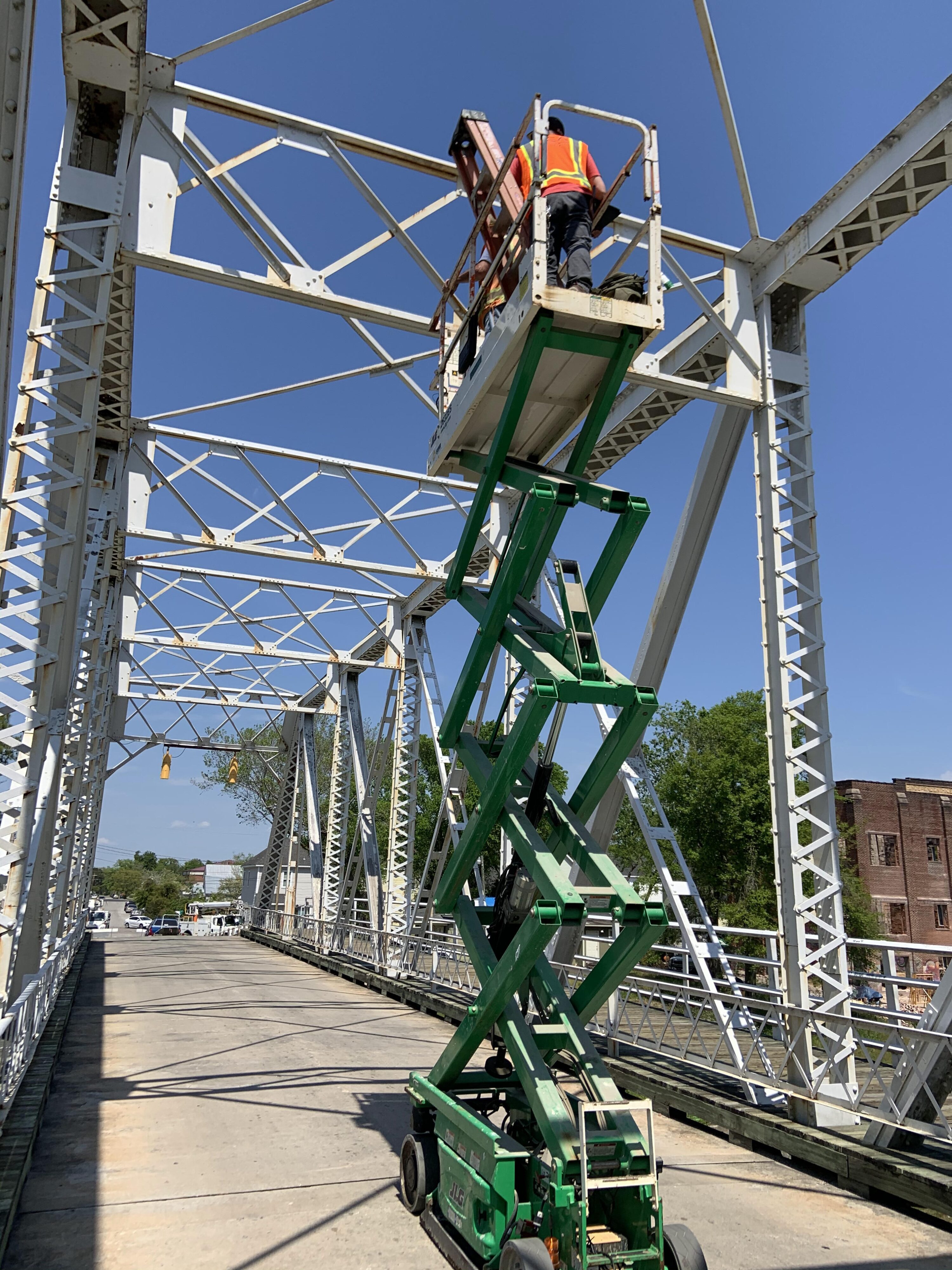 Worker on scissor lift inspecting white steel bridge under clear blue sky