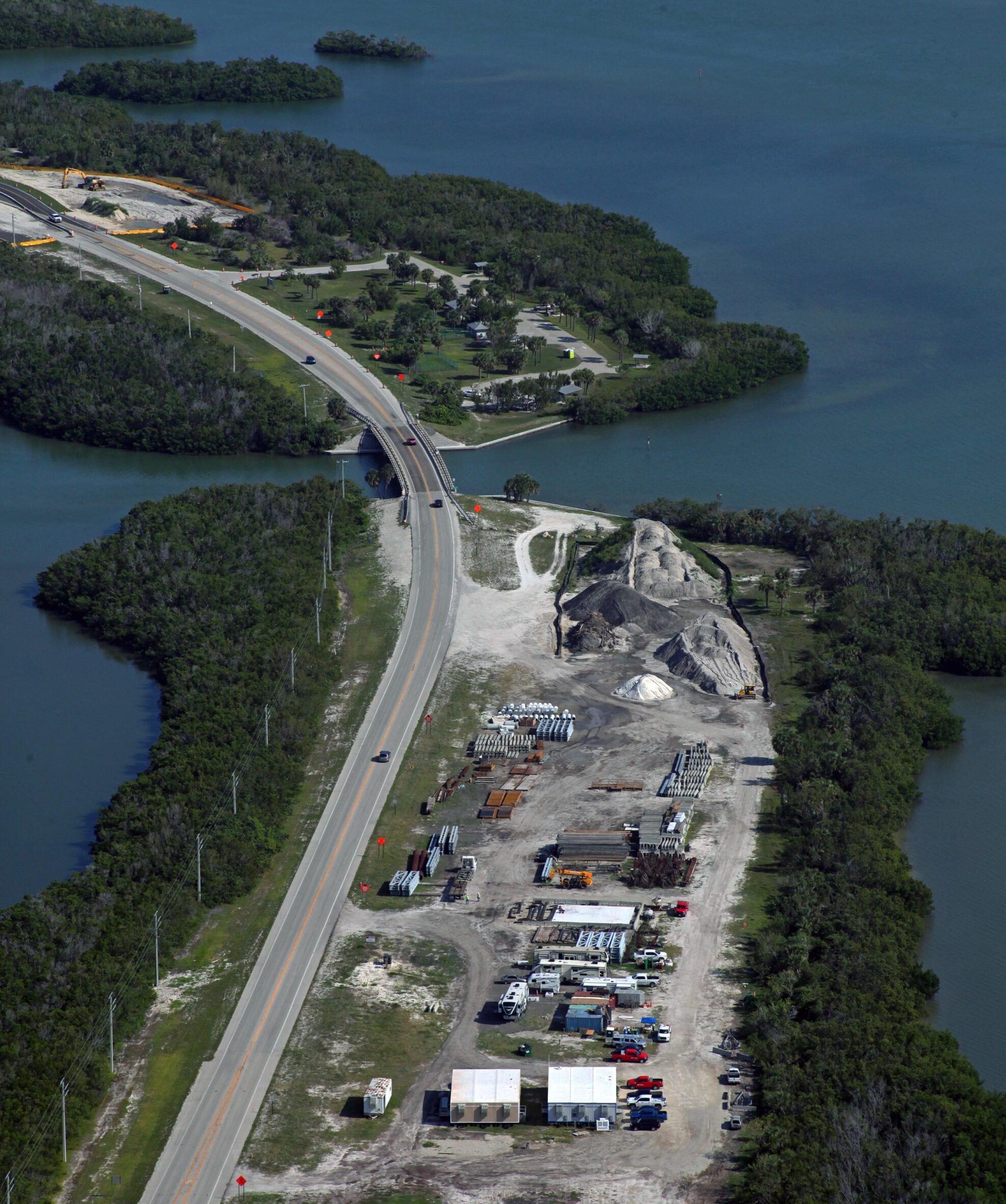 Aerial view of Estero Boulevard with adjacent construction site, surrounded by water and foliage. Signs read: "FAST," "1-800-723-7425," "Florida Aerial Services," "FAST www.FastAerial.com."