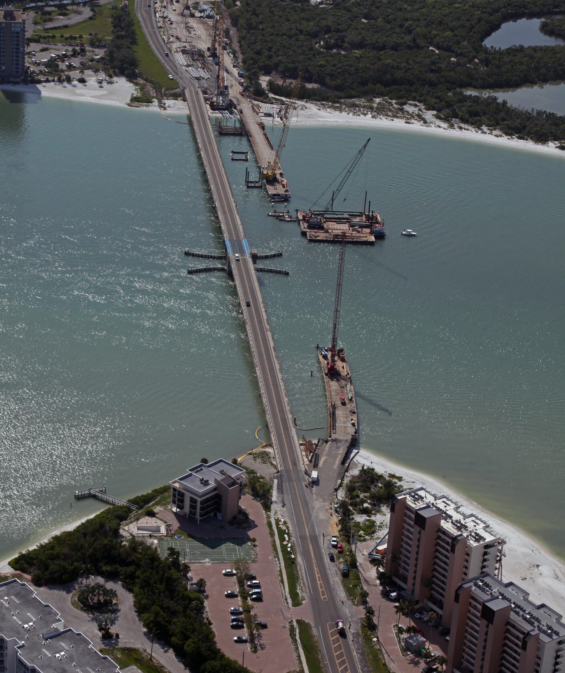 Aerial view of Estero Blvd bridge construction over water, with cranes and adjacent buildings. FAST logo, contact information, and number visible