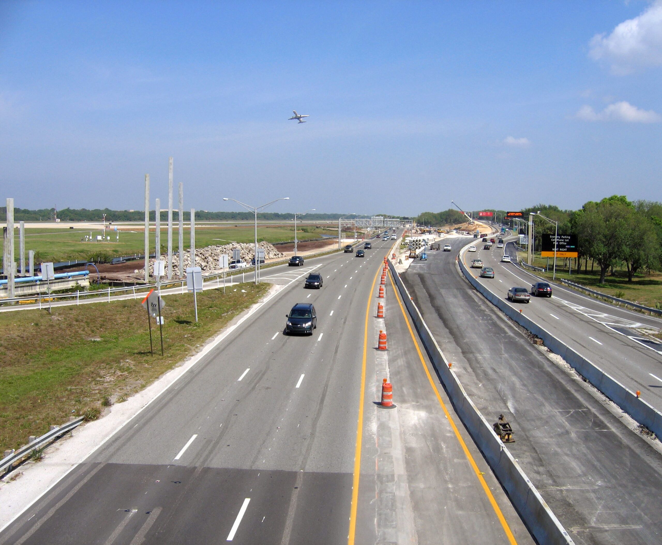Wide highway with vehicles under clear sky, airplane overhead, orange construction cones present