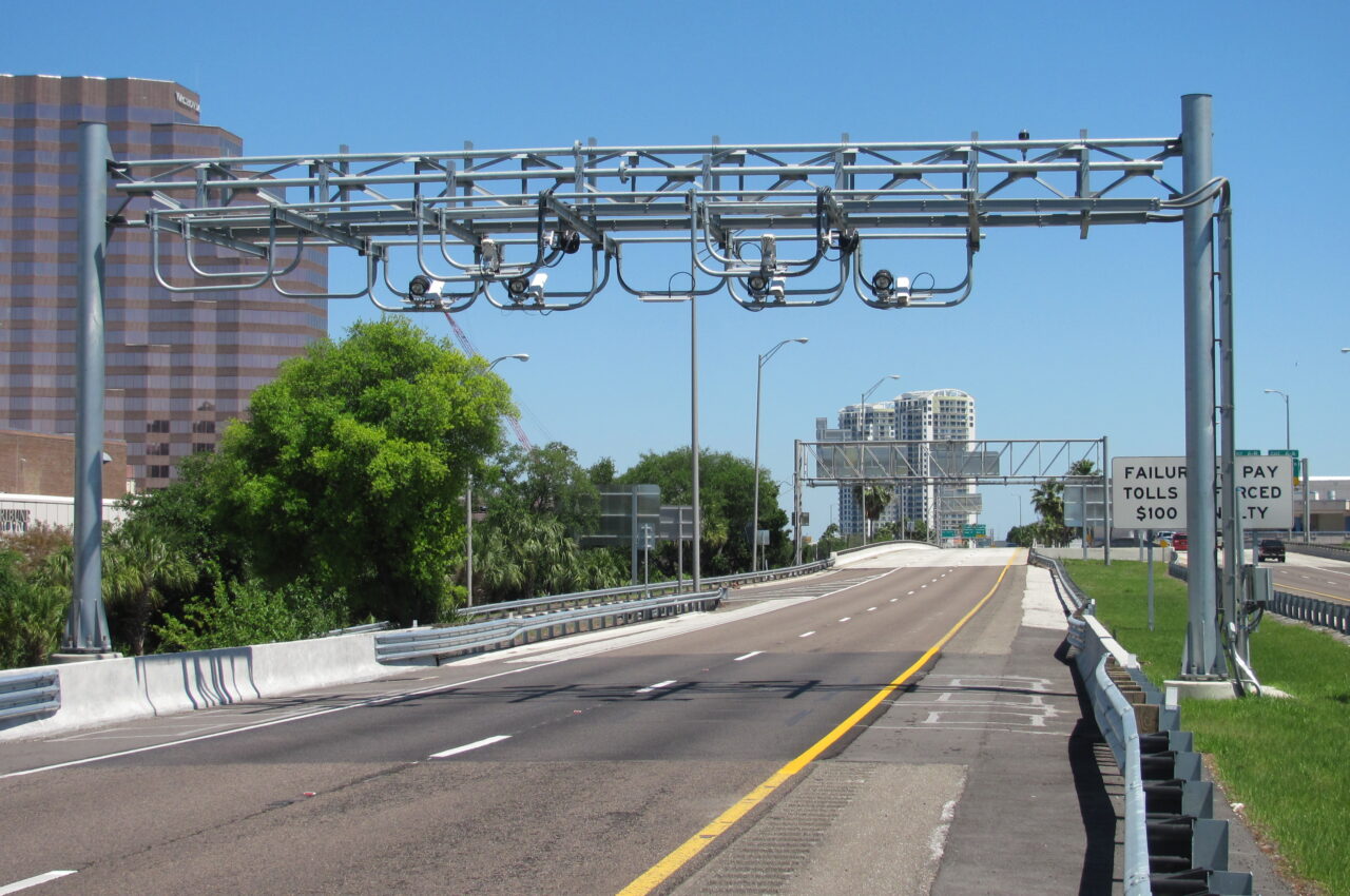 Toll road with electronic gantry, warning signs for unpaid tolls, and city skyline in background