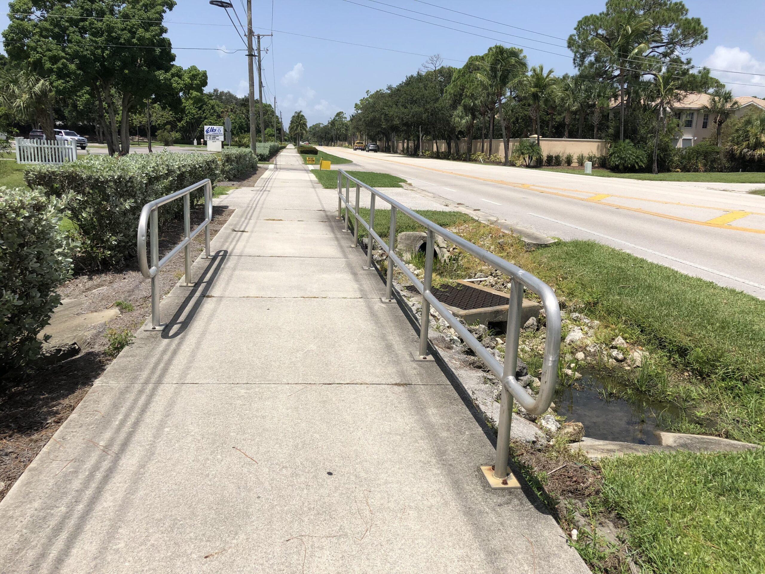 Sidewalk with metal railings, grassy ditch, trees, and a street