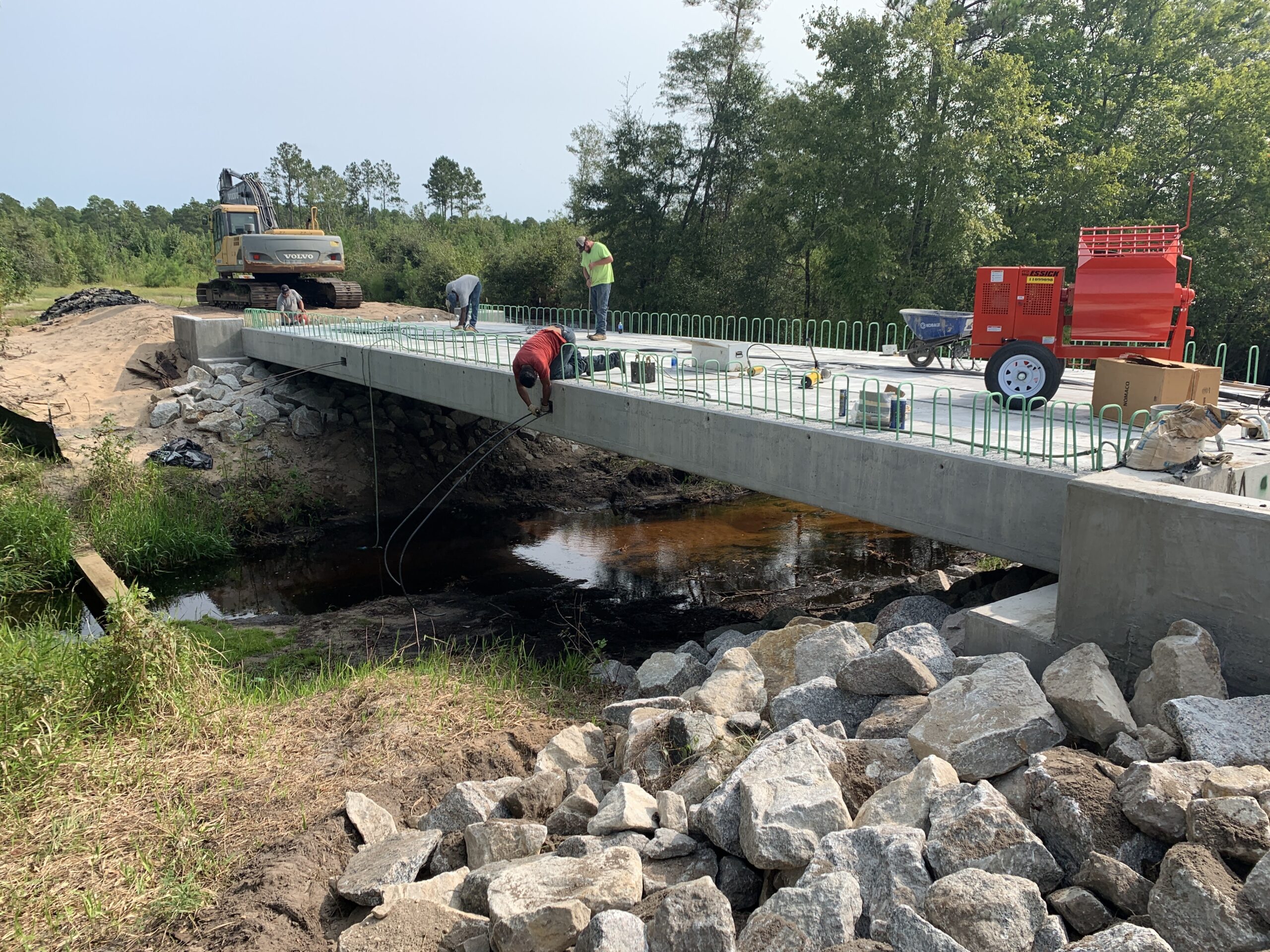 Construction workers on bridge near forested area, operating machinery and laying cables