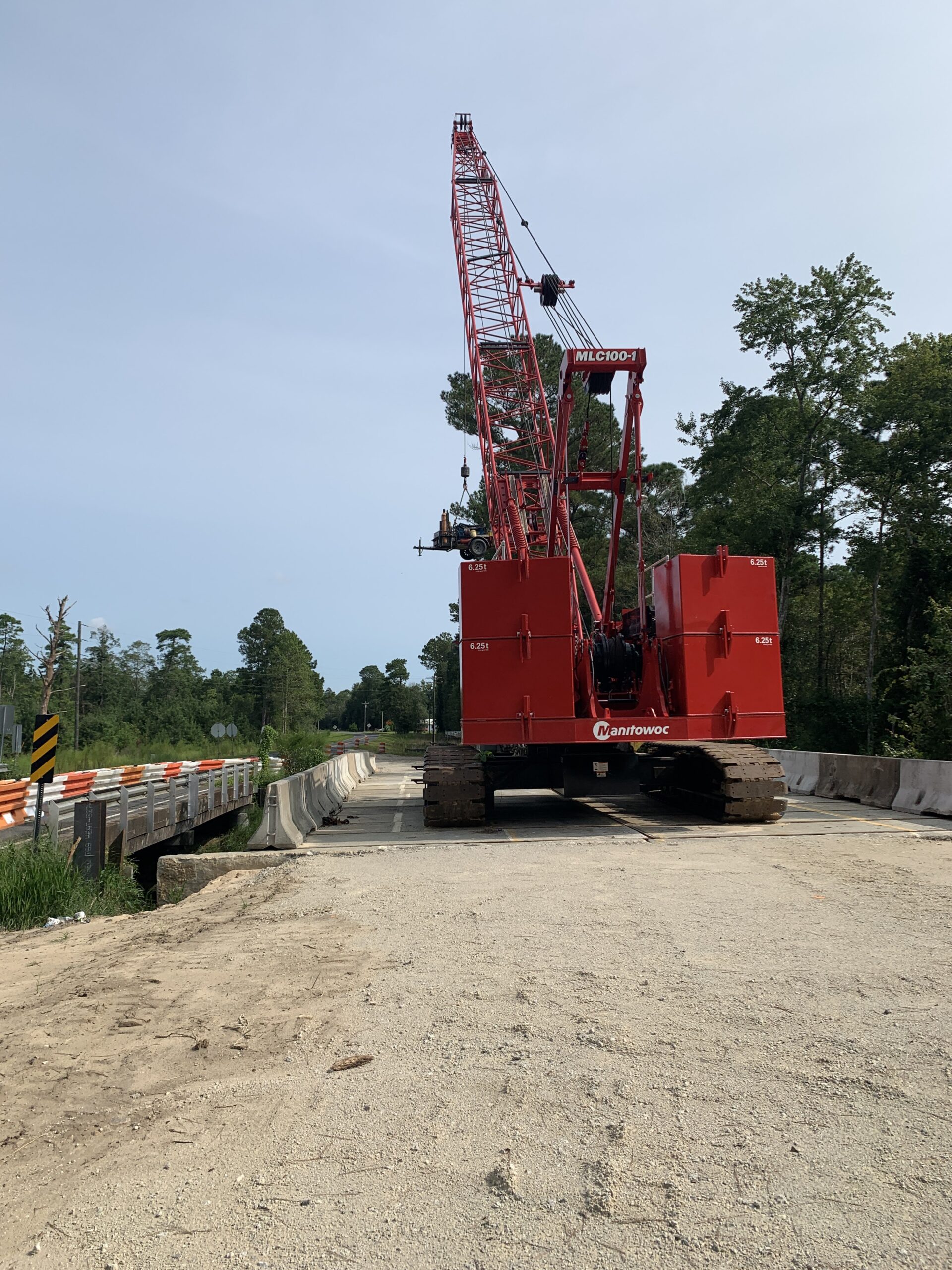Red crane labeled "Manitowoc MLC100-1" on dirt road with trees and barriers