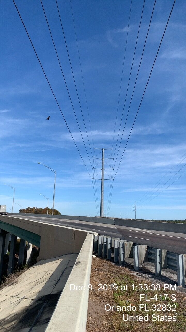 Blue sky above highway overpass with power lines and a distant bird. Text: "Dec 9, 2019 at 1:33:38 PM, FL-417 S, Orlando FL 32832, United States" in white text on image