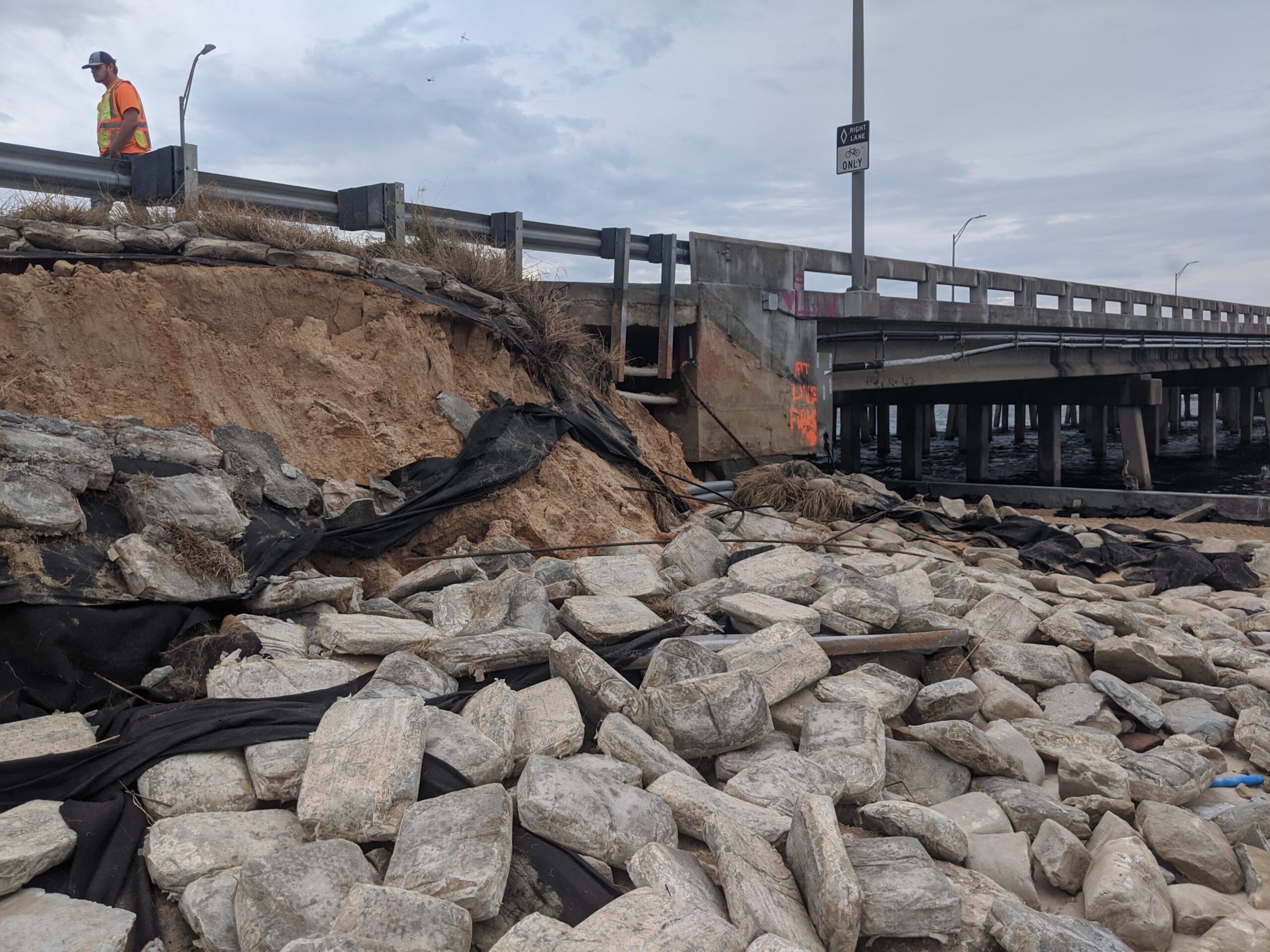 Eroded bridge embankment with exposed rocks and geotextile, worker inspecting damage