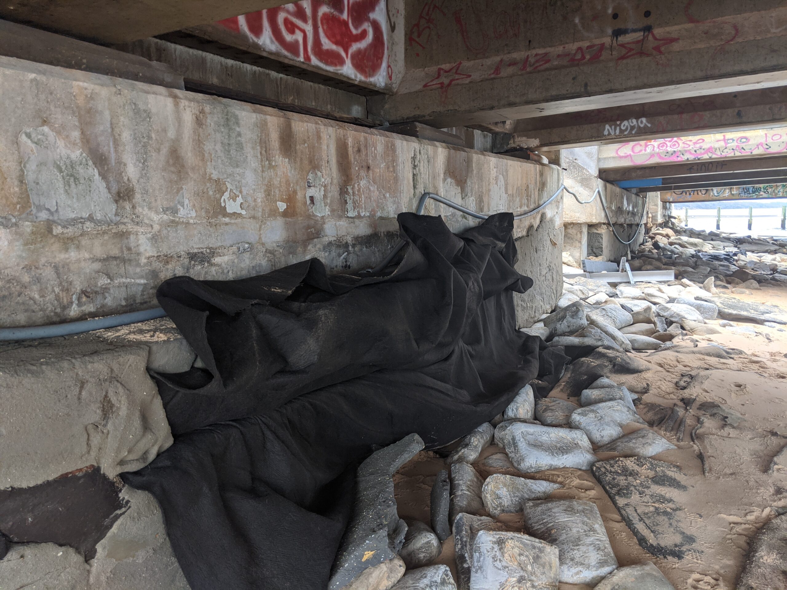 Underpass with graffiti-covered concrete and black tarp, rocks scattered