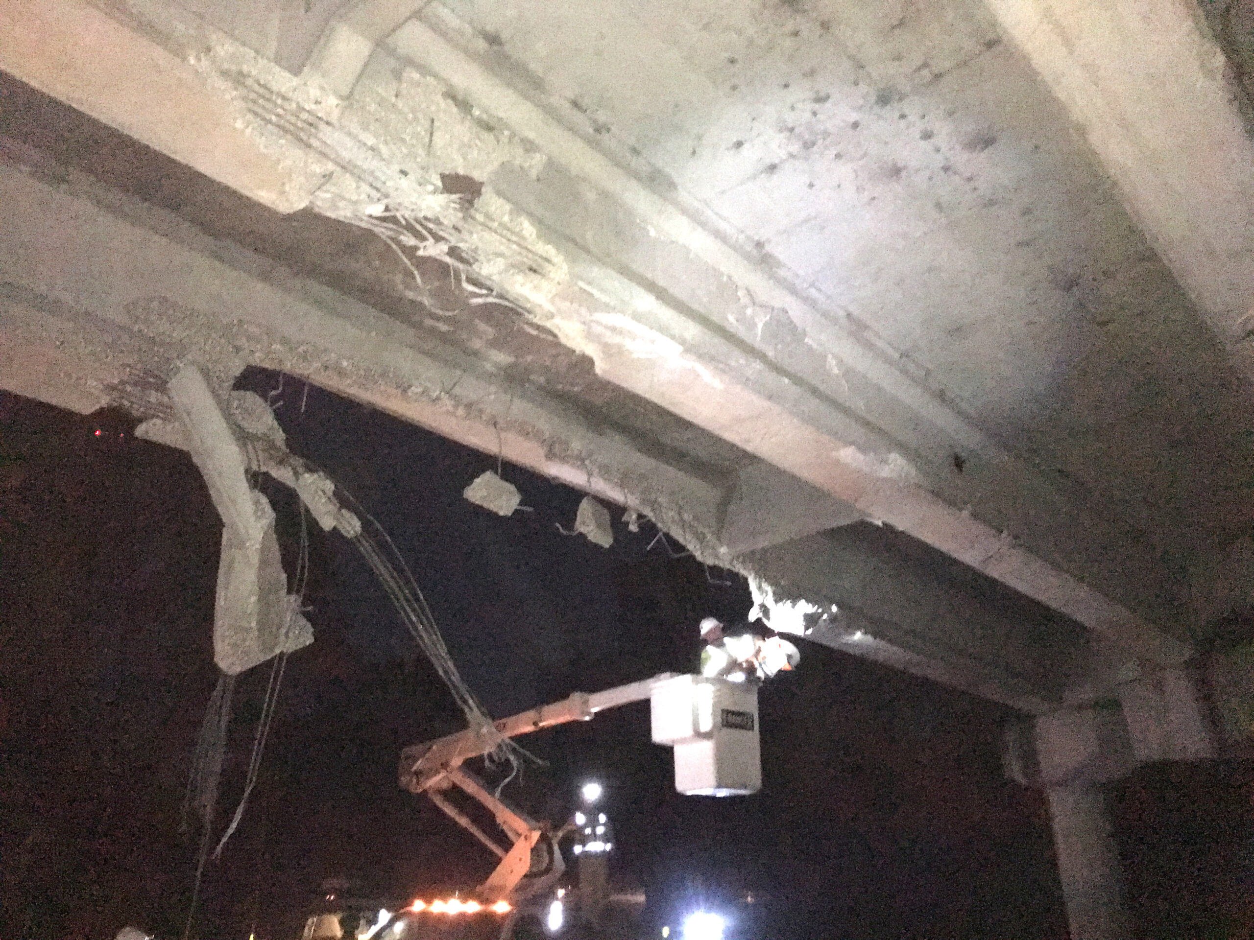 Damaged bridge undercarriage with exposed rebar, workers in lift inspecting at night