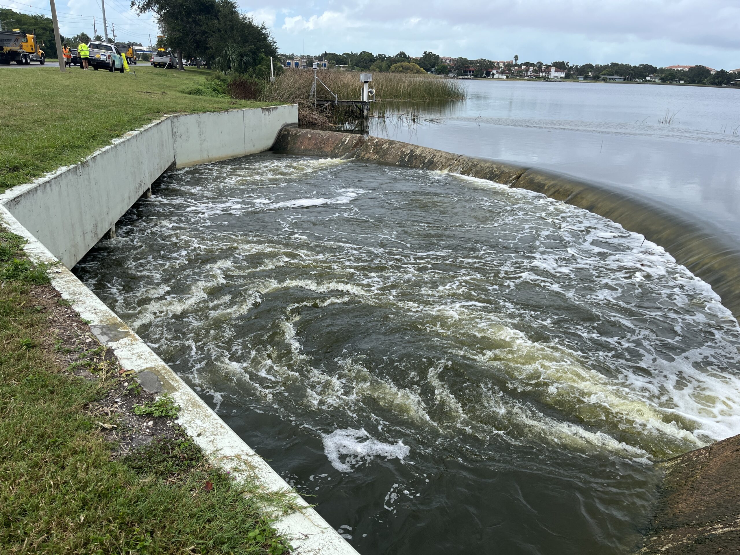 Grass-lined drainage area with water flowing over a weir, utility workers nearby