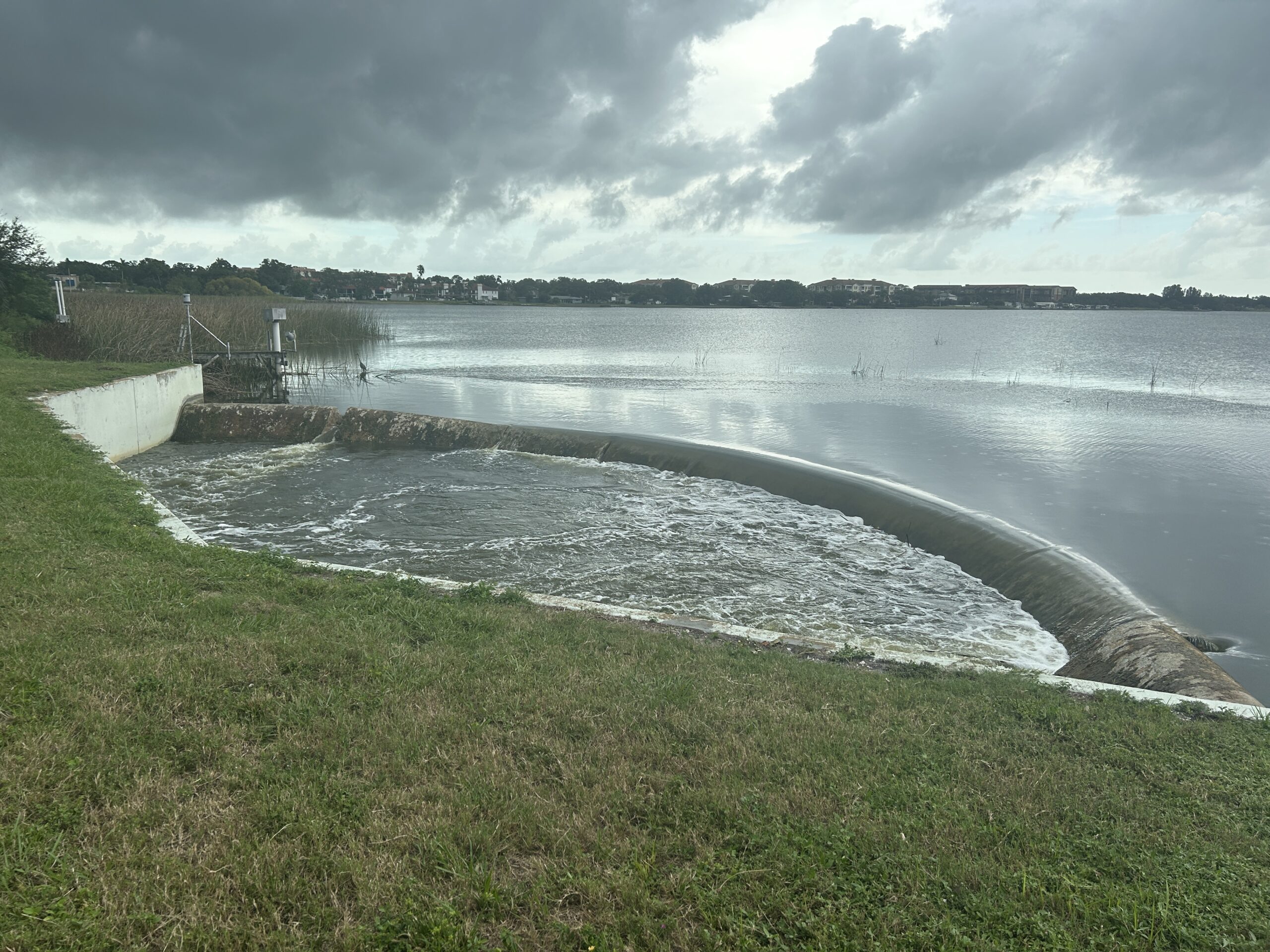 Water flowing over a curved dam into a lake under cloudy skies