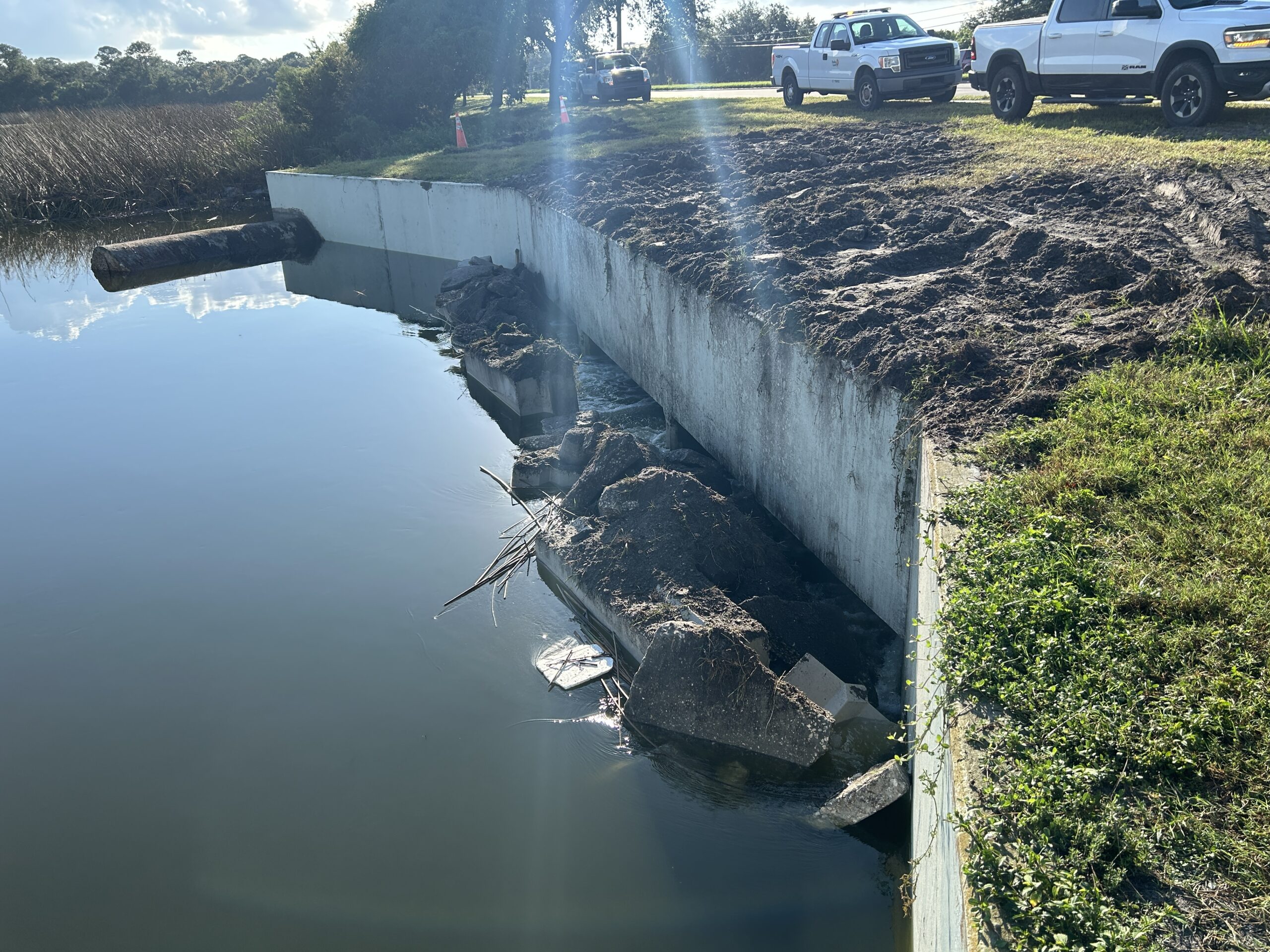 Damaged concrete embankment with scattered debris, adjacent to a calm water body, flanked by parked trucks and grassy terrain