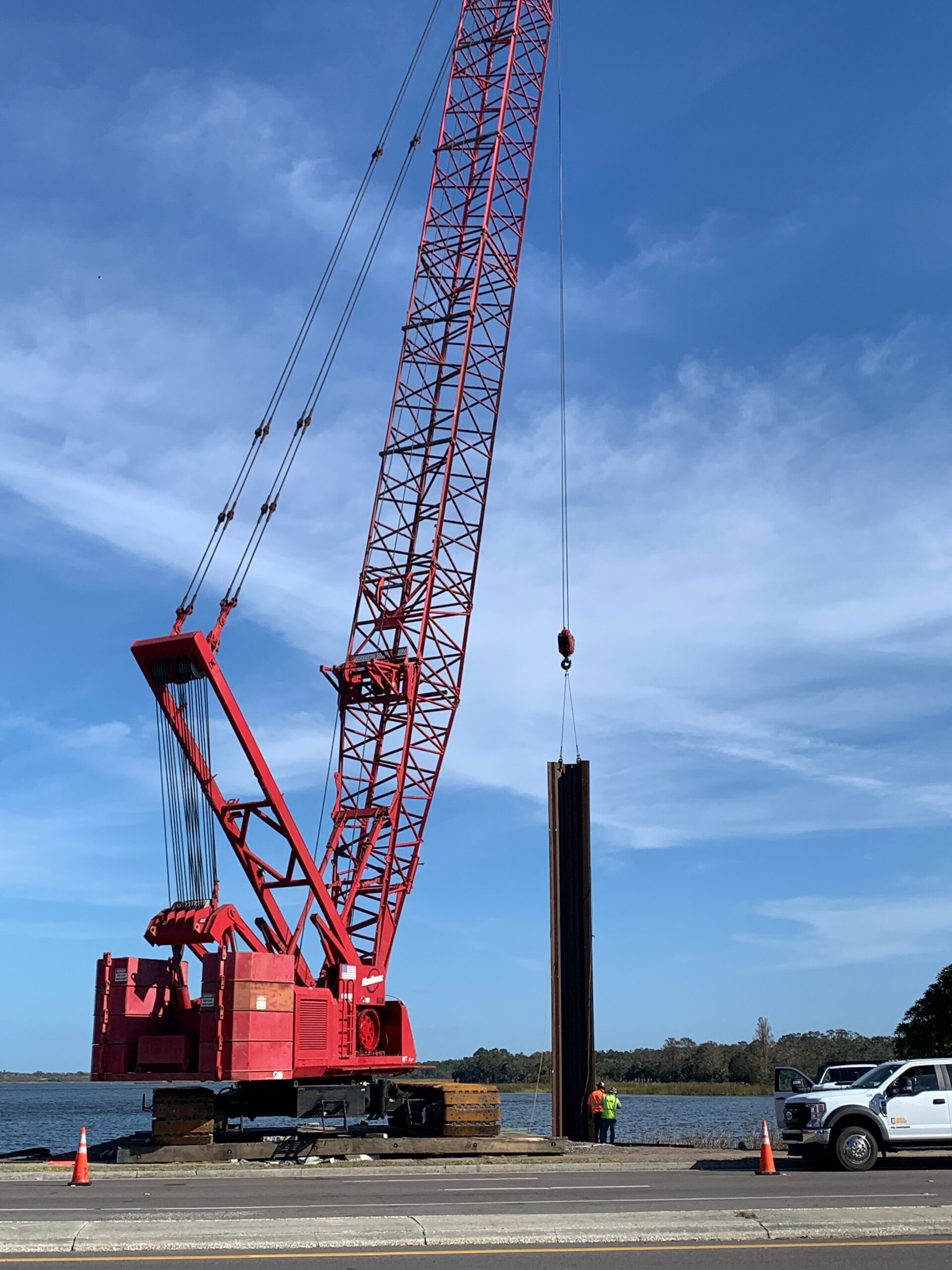 Red construction crane lifting steel beams near water, two workers nearby