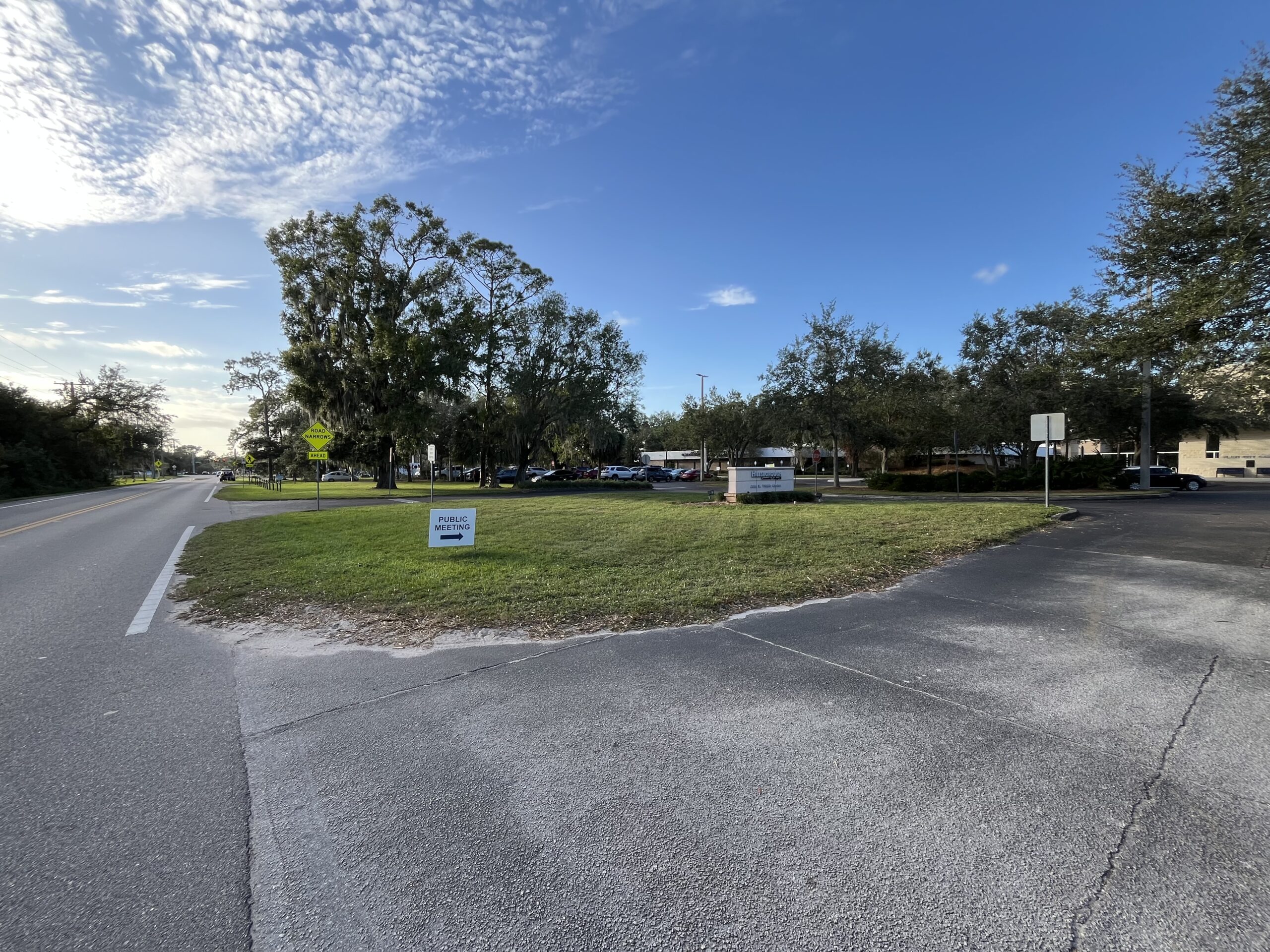 Public meeting sign on grass by road with trees and parking lot in background