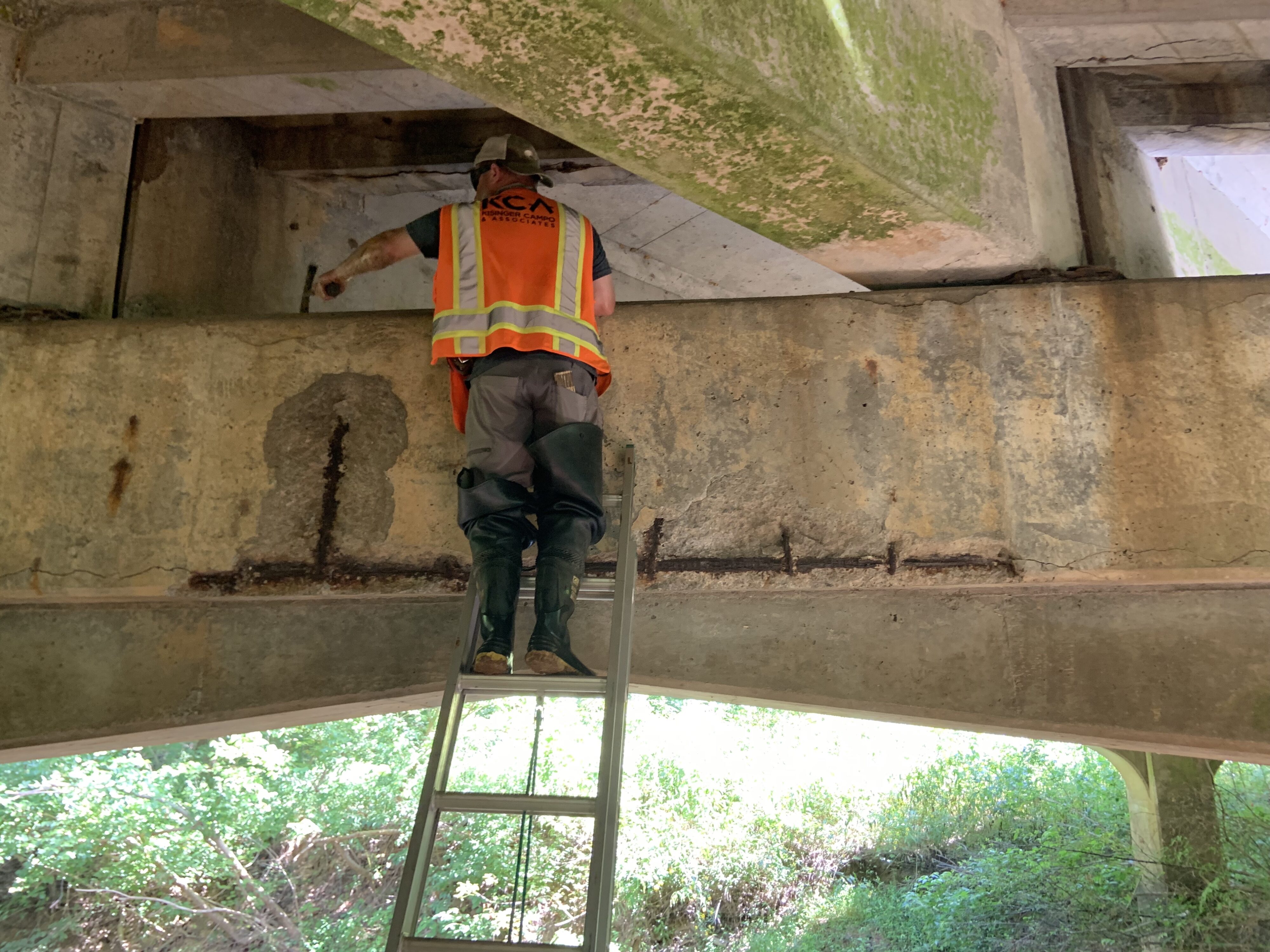 Man on ladder inspecting bridge underside, wearing safety gear and orange vest