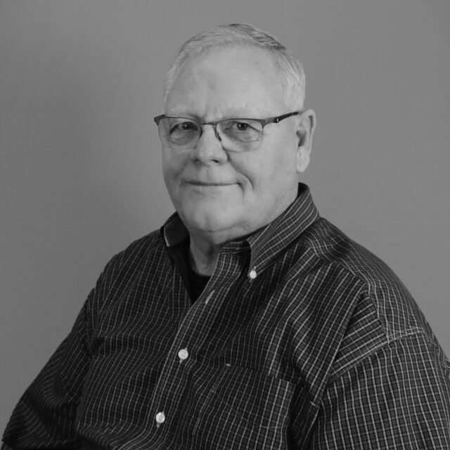 Kays-Bill smiling in a black and white portrait, wearing glasses and a checkered shirt