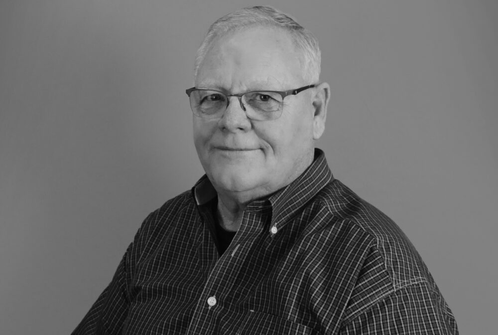 Kays smiling in black and white, wearing glasses and a checkered shirt