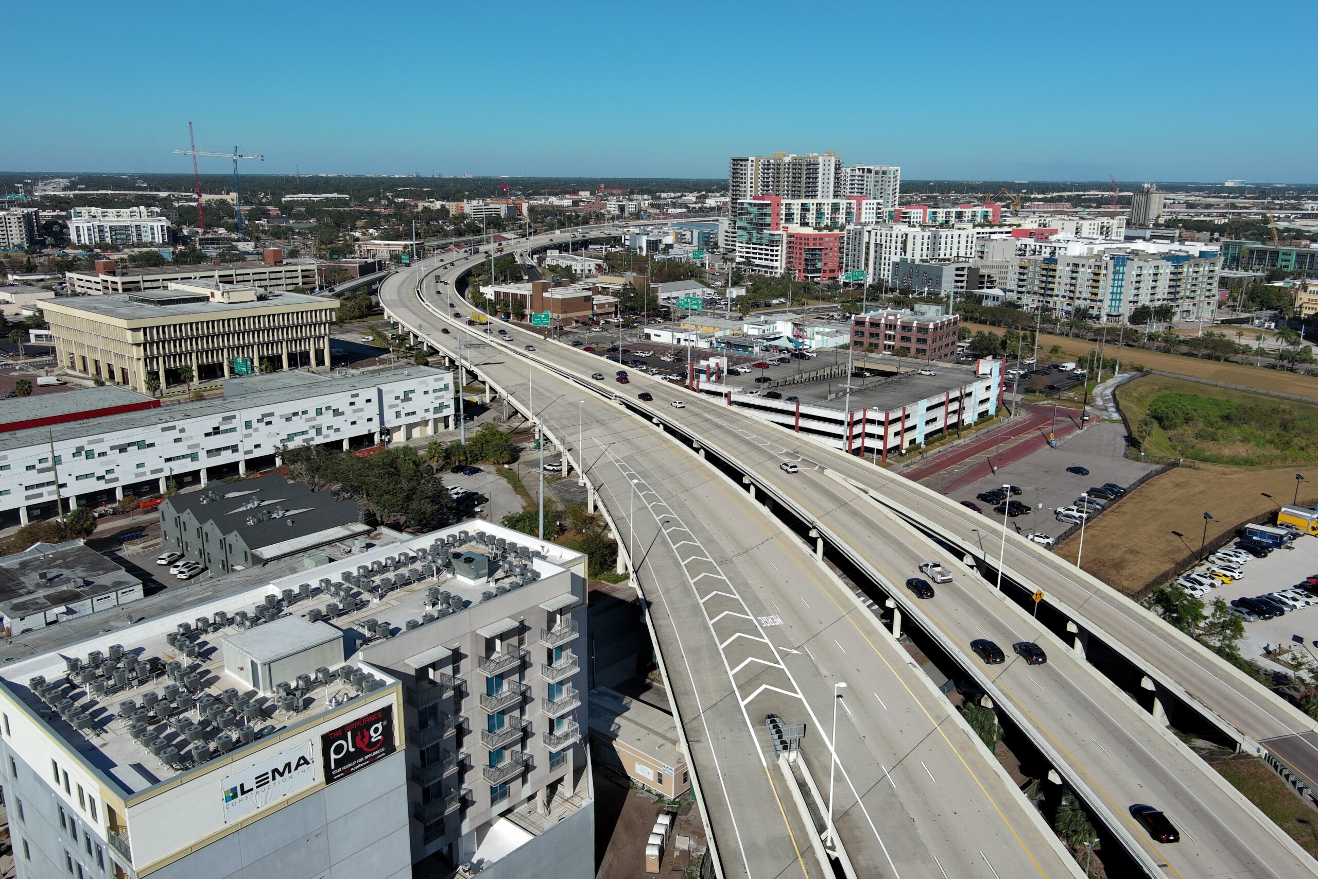 Aerial view of a highway overpass through a cityscape with buildings and construction cranes, Lema and The Plug signs visible