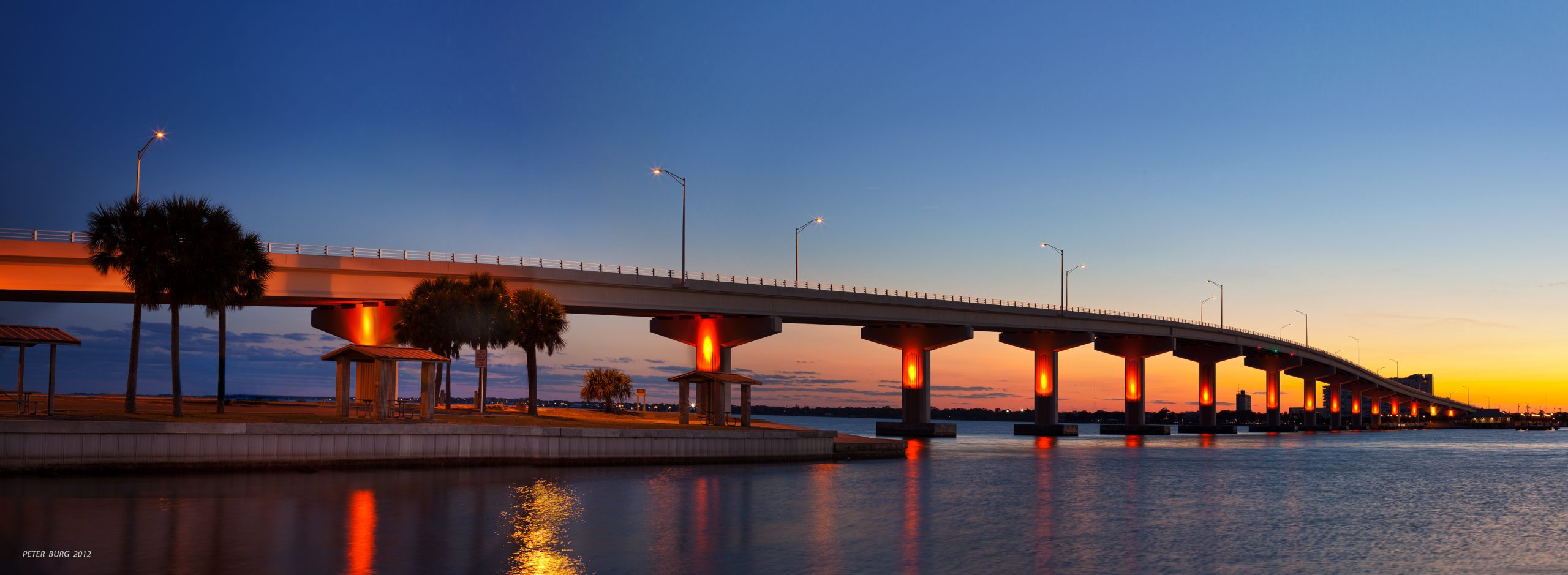Bridge over water at sunset, orange lights illuminate underside, palm trees nearby