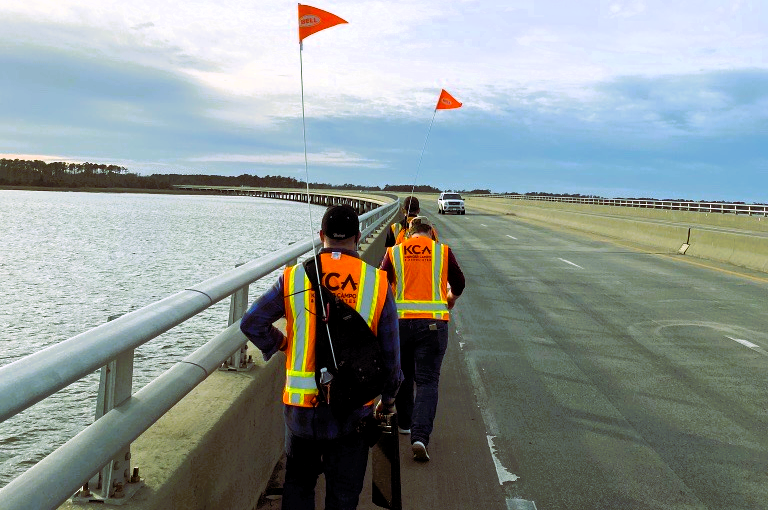 Two workers in safety vests walk along bridge, carrying gear, with water beside them