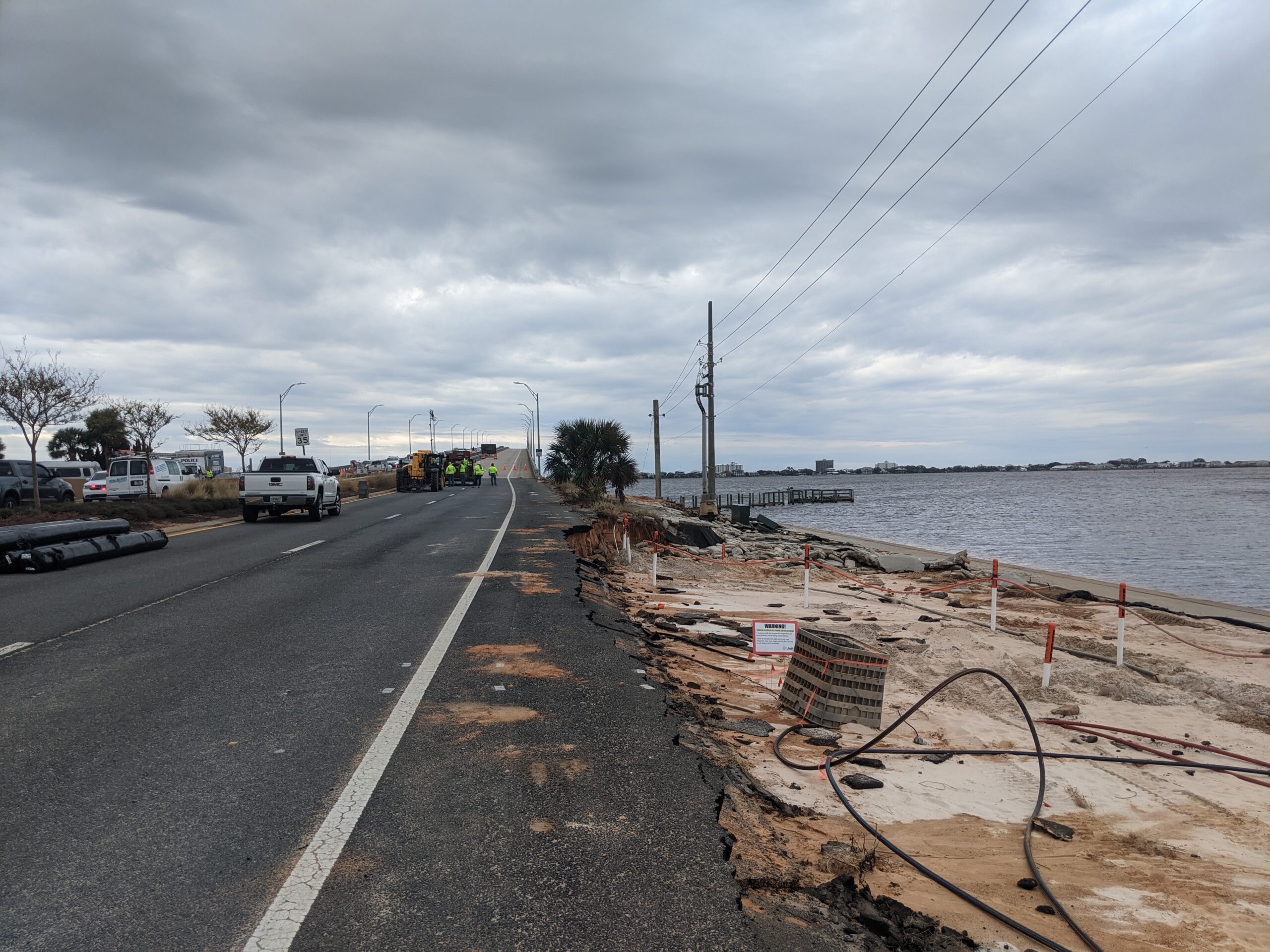 Road construction along waterfront with heavy machinery and workers repairing eroded section