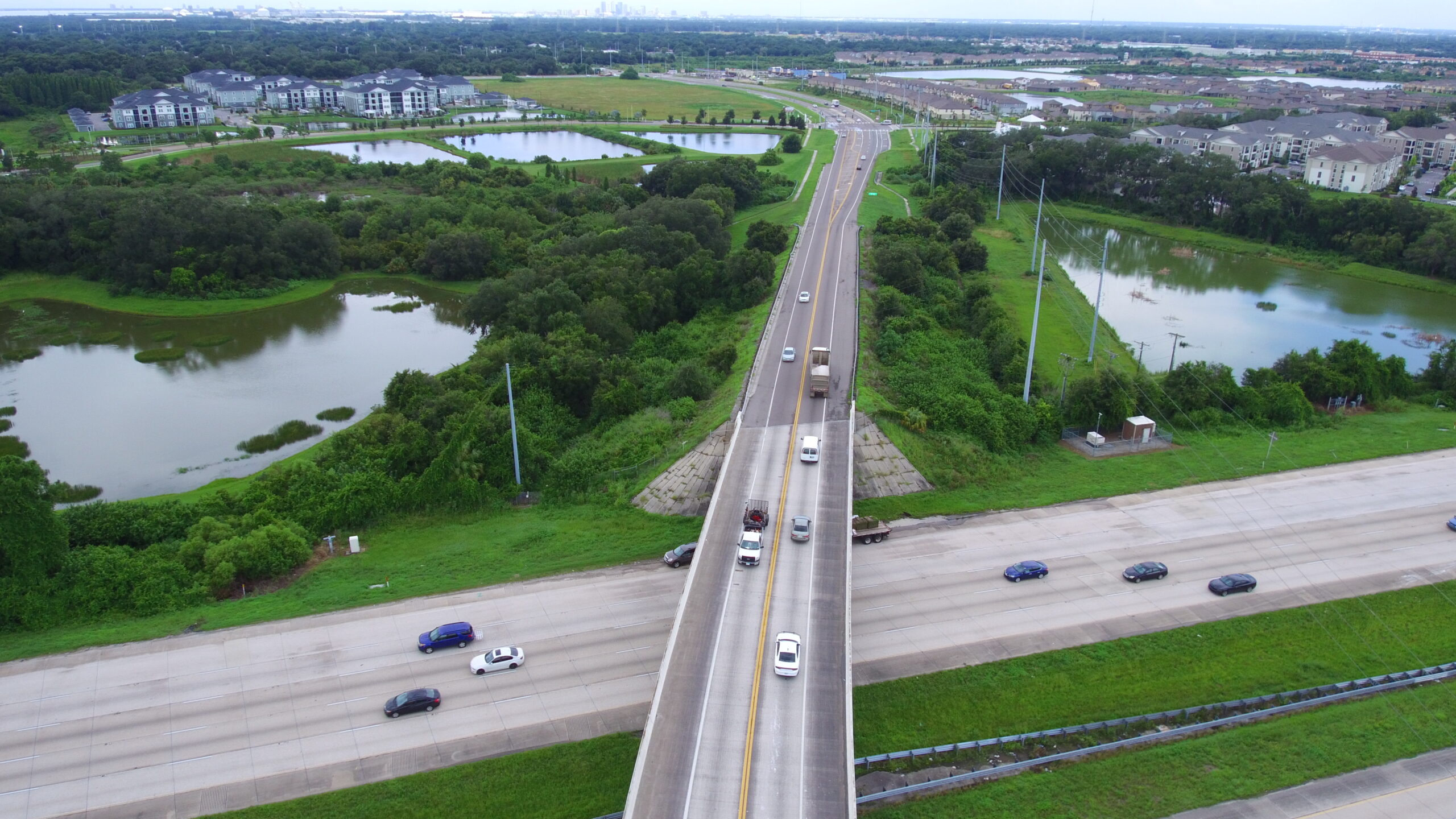 Aerial view of a highway bridge over a forested area with surrounding ponds and residential buildings in the distance