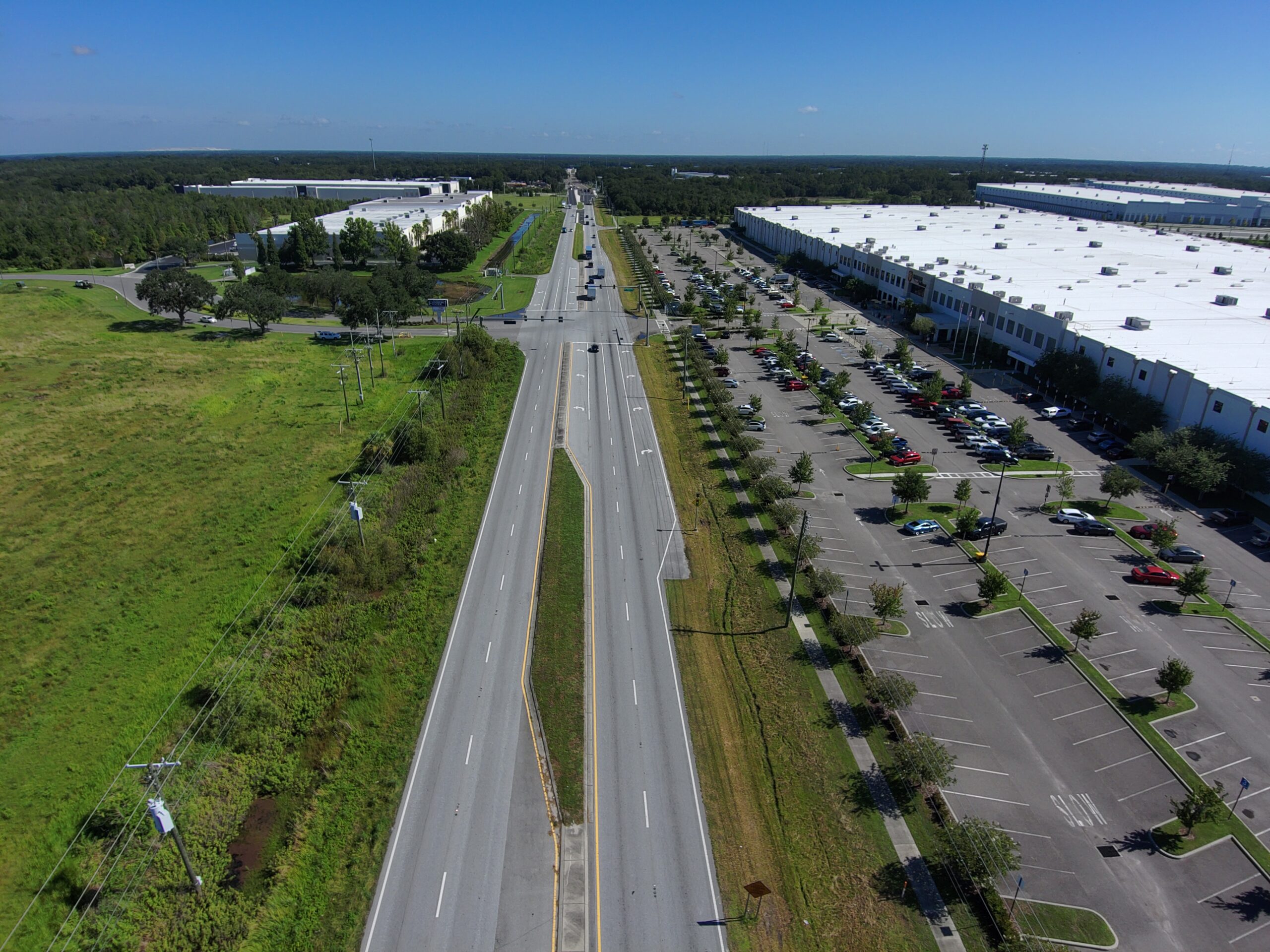 Aerial view of a wide, empty road flanked by grassy areas and a parking lot with a large white-roofed warehouse adjacent