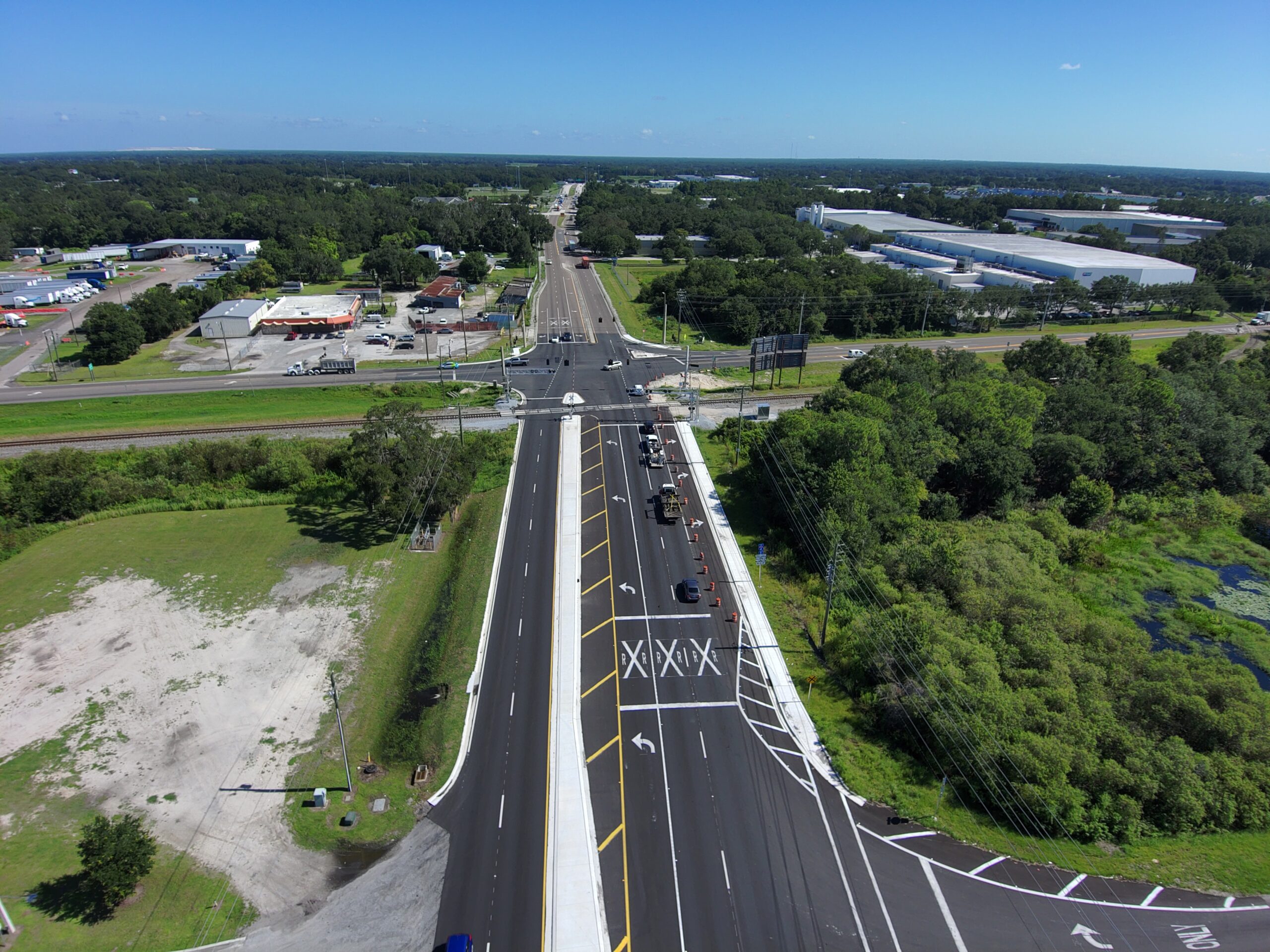 Aerial view of a multi-lane highway intersection, surrounded by greenery and industrial buildings in the distance under a clear blue sky