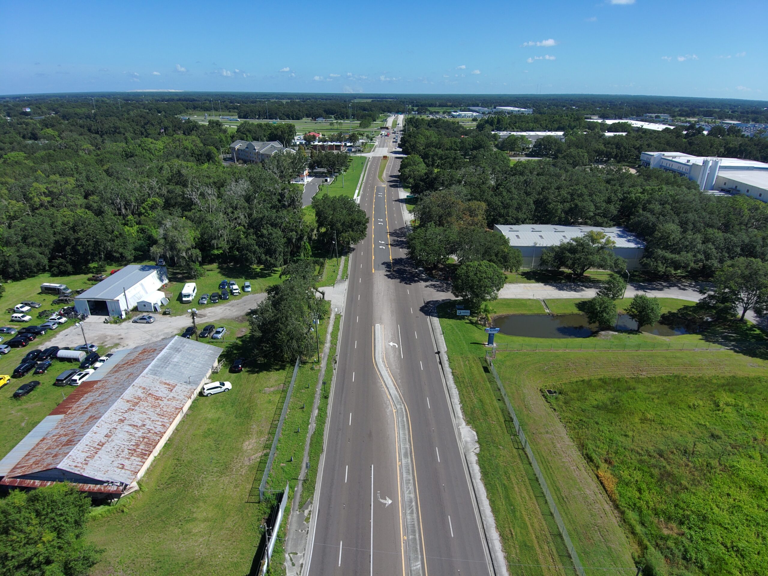 Aerial view of a two-lane road flanked by green trees and industrial buildings on a clear, sunny day