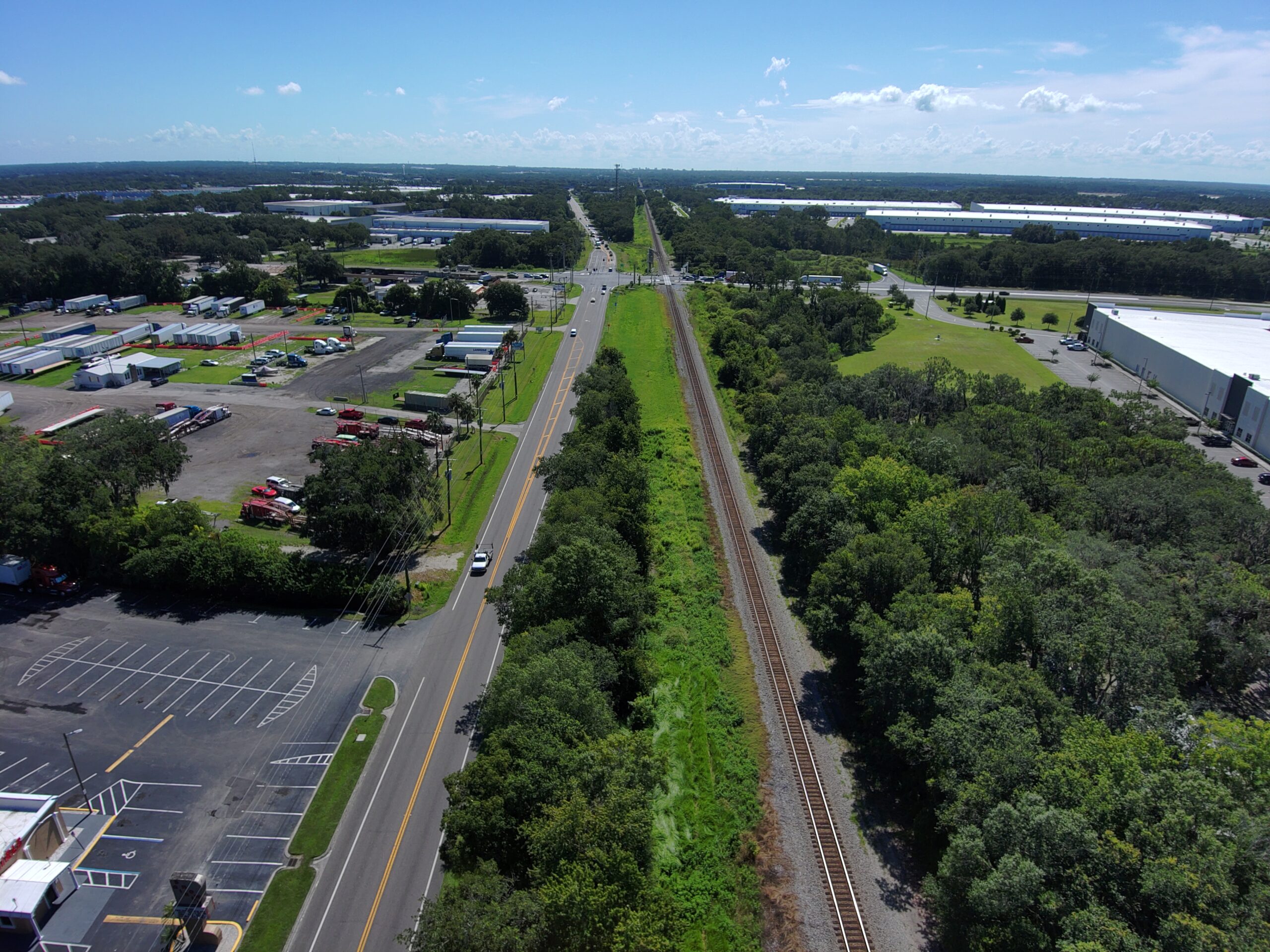 Aerial view of a road beside railroad tracks, industrial buildings, and greenery under a clear blue sky