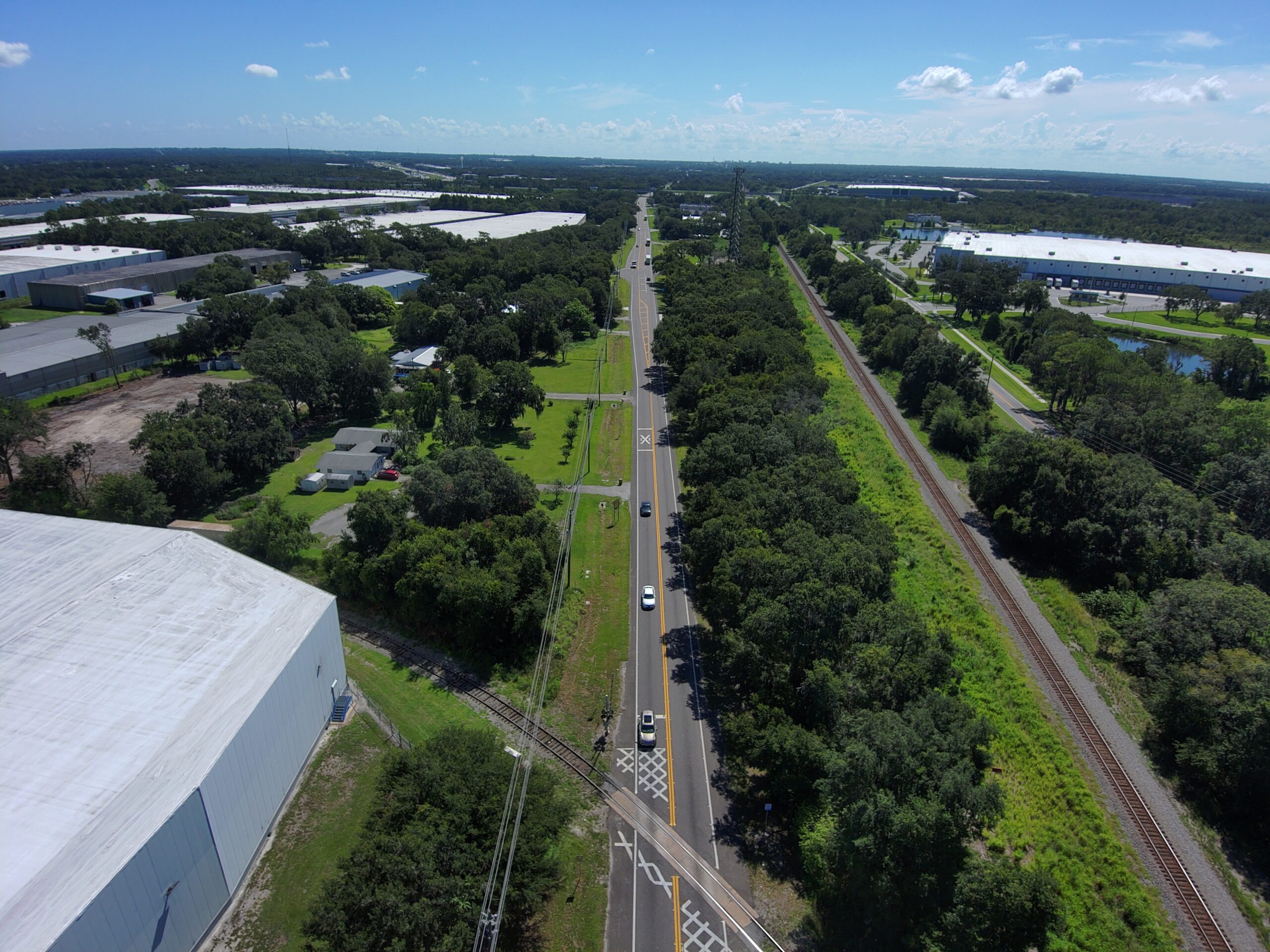 Aerial view of a straight road flanked by warehouses and lush green trees, with a railway line on the right