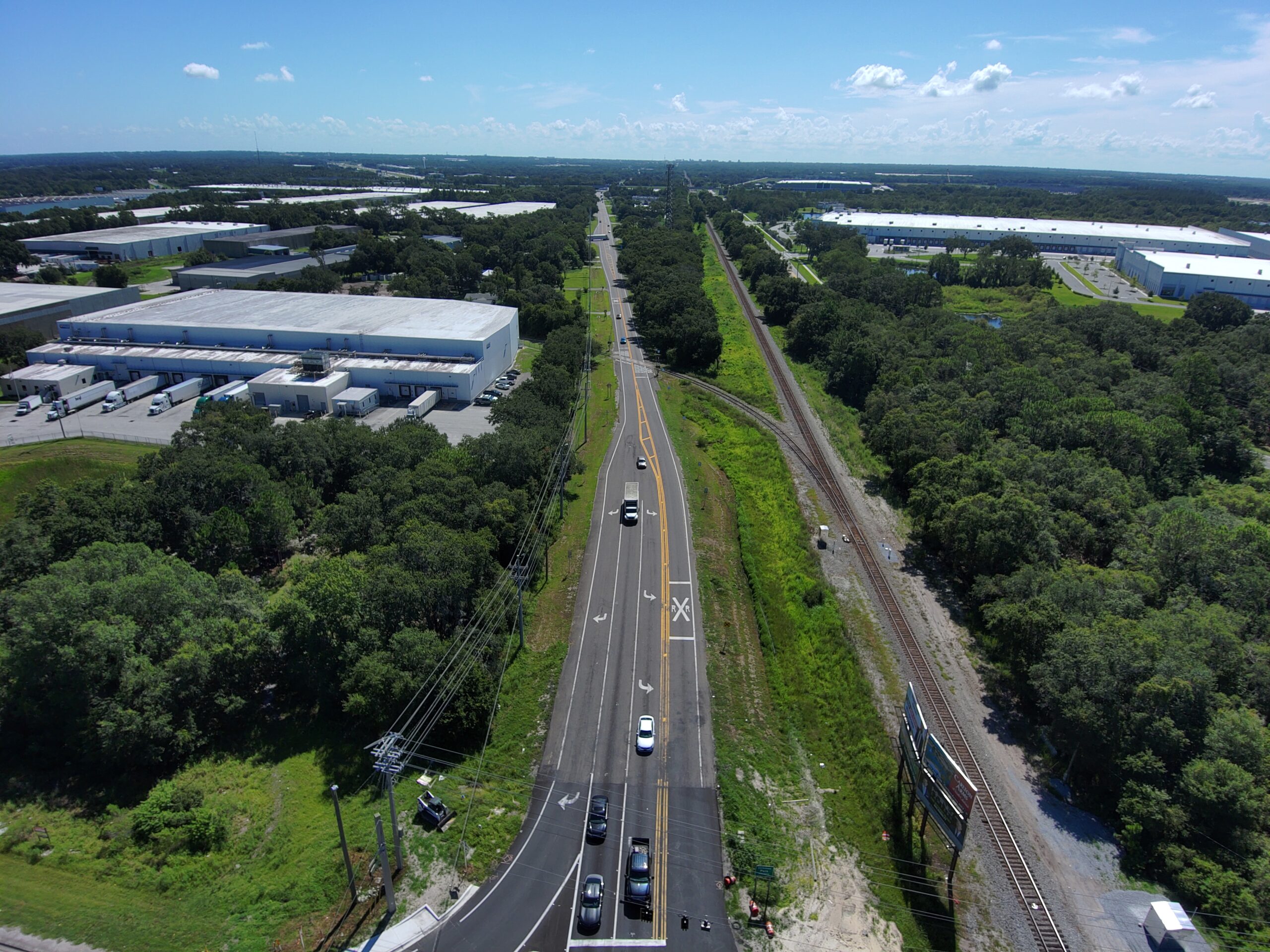Aerial view of a road with vehicles, adjacent to a railway track and industrial buildings surrounded by trees