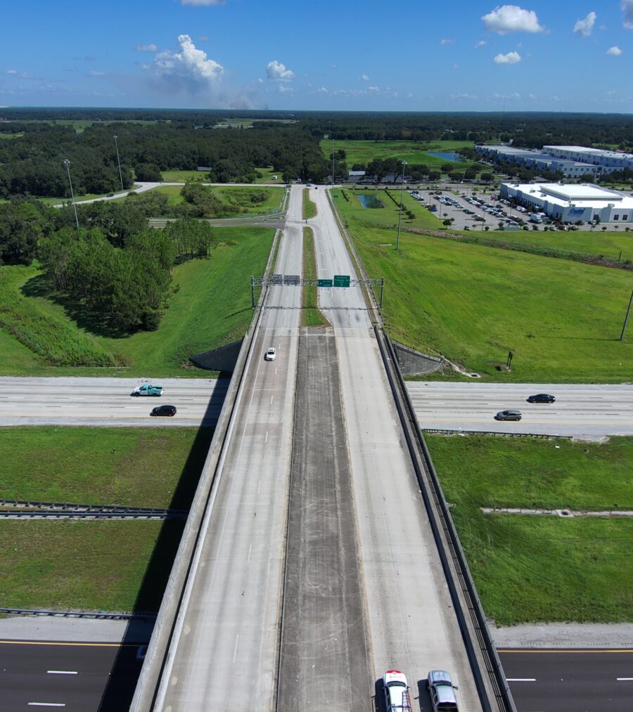 Elevated highway intersection with light traffic, surrounded by green fields and distant industrial buildings under a blue sky
