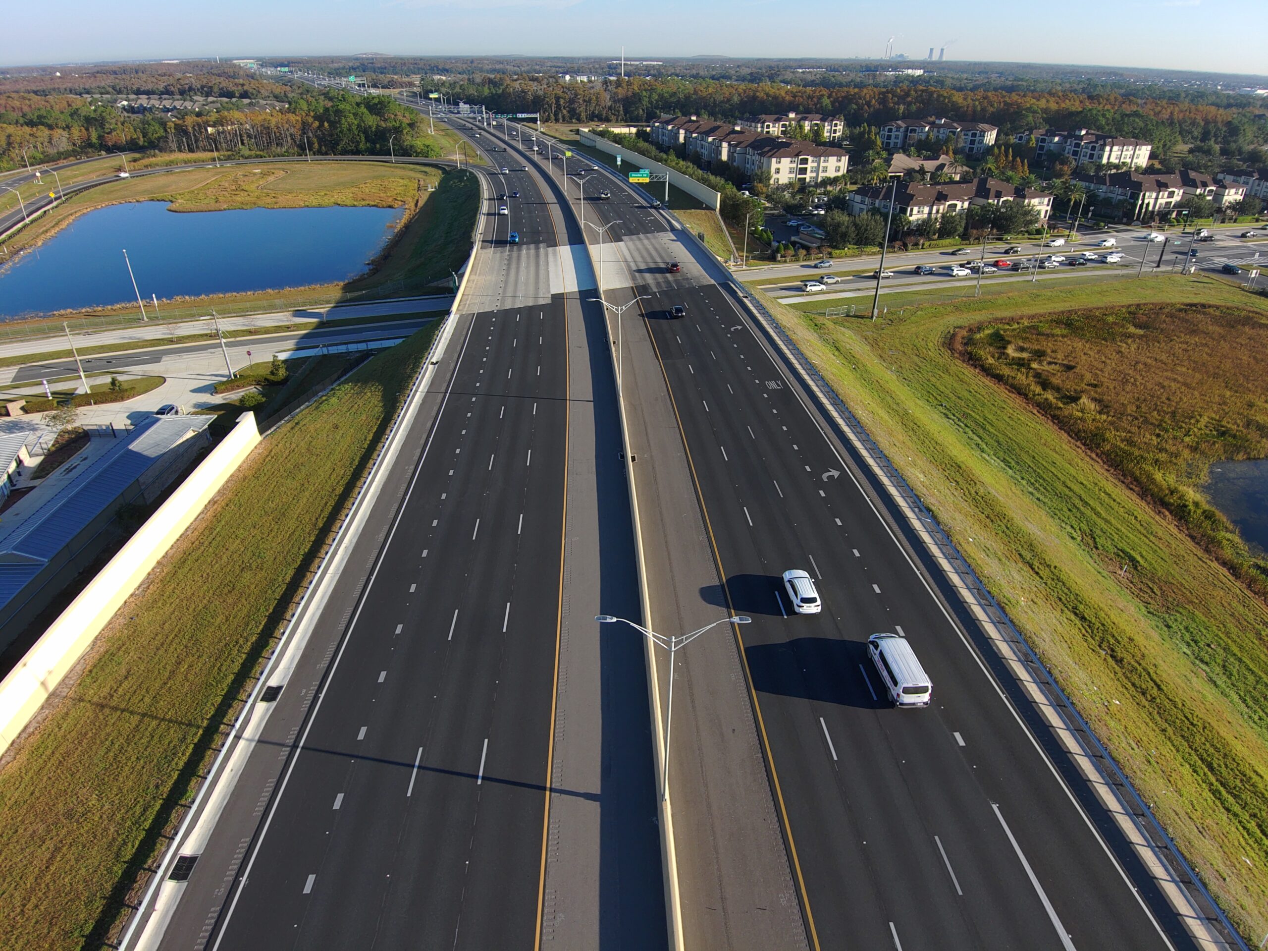 Aerial view of a wide, empty highway with sparse traffic, flanked by greenery, residential area, and a pond
