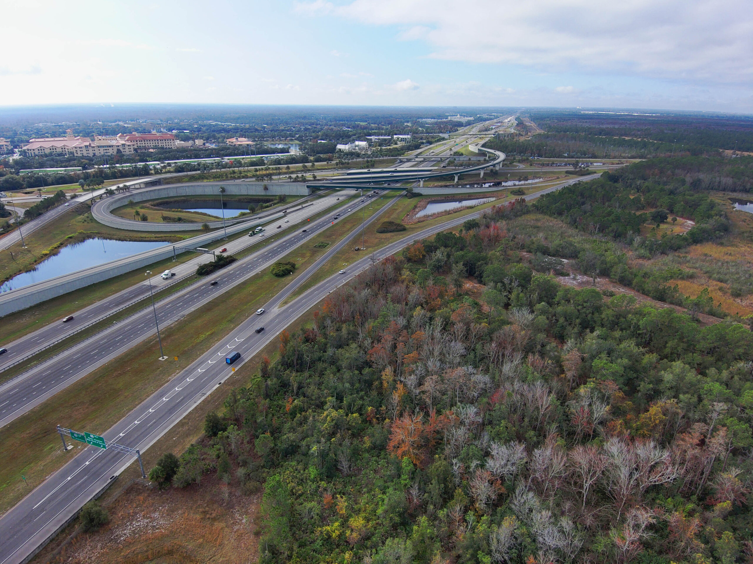 Aerial view of a highway interchange with looping overpasses, surrounded by forests, ponds, and urban structures in the background