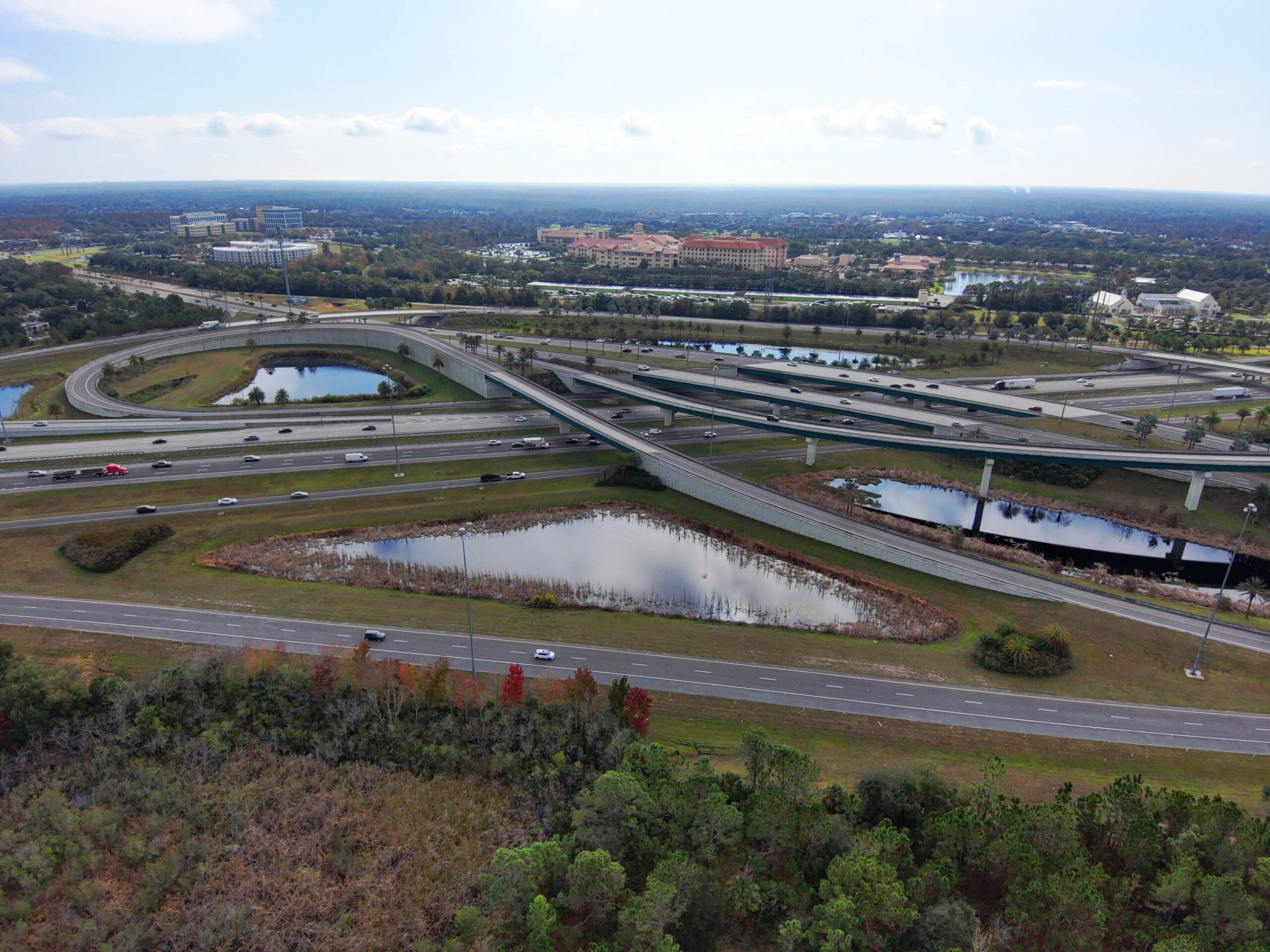 Aerial view of multi-lane highway interchange with surrounding ponds and greenery, under a partly cloudy sky