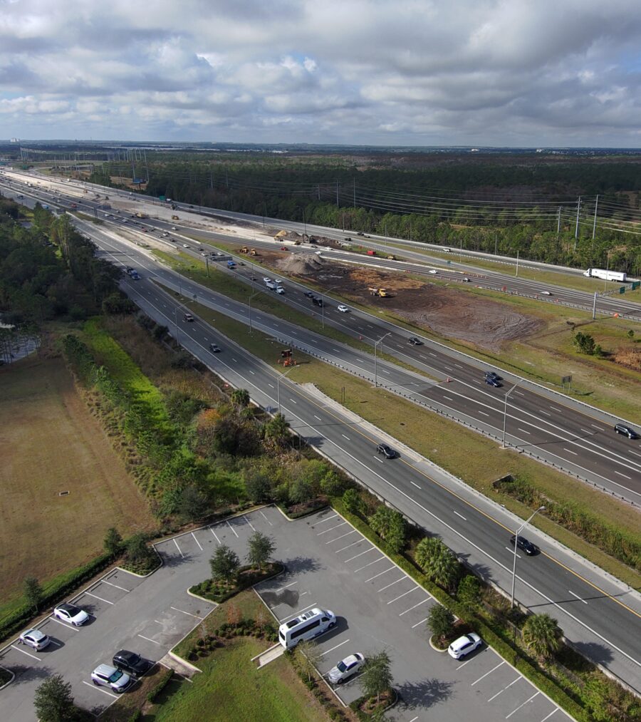 Highway construction scene with scattered vehicles and adjacent greenery under a cloudy sky