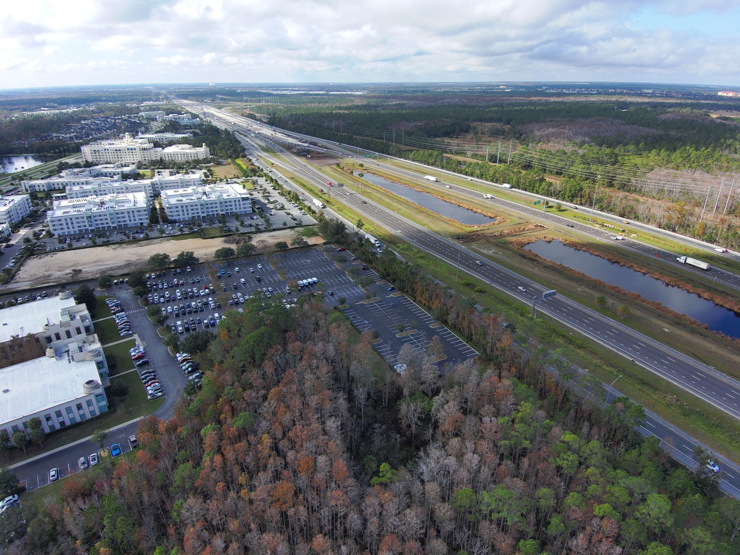 Aerial view of a highway beside a commercial area and dense forestland, under a cloudy sky