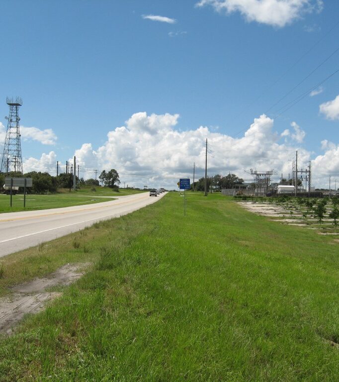 Highway with lush greenery, transmission tower, and industrial site under a sunny sky