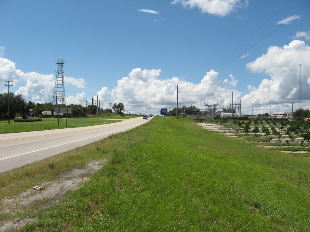 Highway with lush greenery, transmission tower, and industrial site under a sunny sky