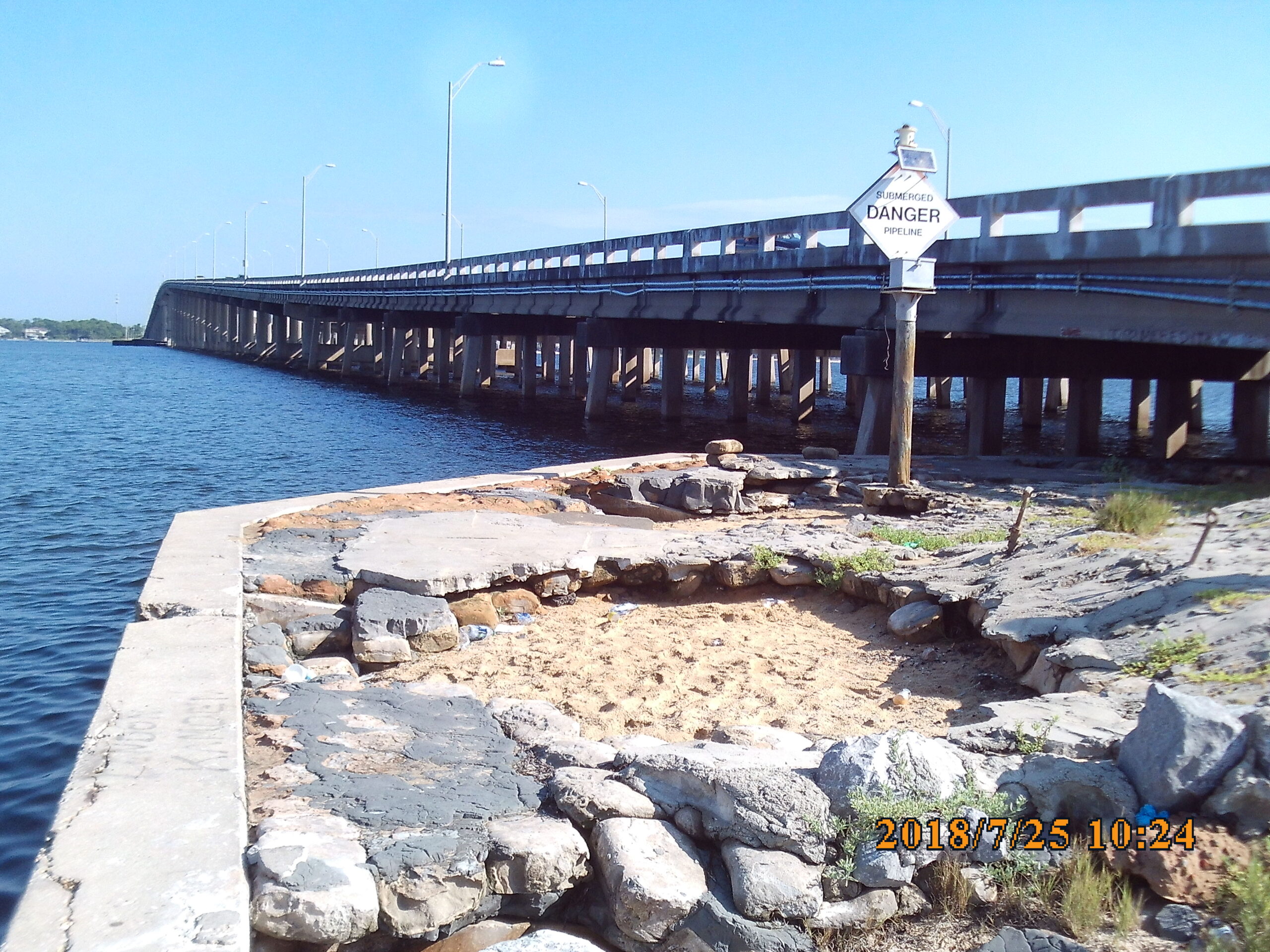 Bridge over water with "Submerged Danger Pipeline" sign visible. Rocky shore in foreground, date displayed: 2018/7/25 10:24